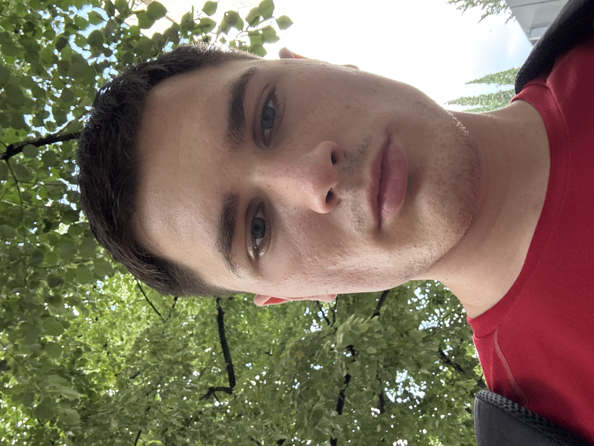 A close-up, low-angle, selfie-style photograph features a young Caucasian man with short, dark hair, a light complexion, and a neutral facial expression. He is wearing a red t-shirt and a dark backpack strap is visible over his left shoulder. The background is dominated by lush green foliage, suggesting an outdoor setting with trees. A bright, overcast sky is visible in the upper right portion of the image, with some modern buildings and tall, slender trees in the distance on the right. The lighting is bright and diffused, casting soft shadows. The composition focuses on the man's face and upper torso, with the natural elements framing him. There is no identifiable context to place the image specifically within Stara Zagora, Bulgaria, beyond the provided information.