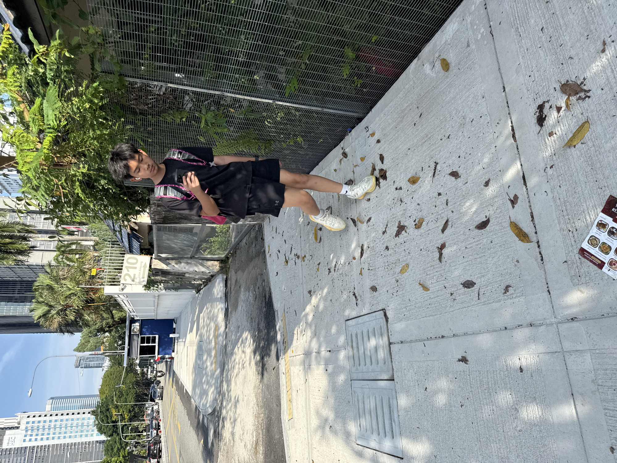 An eye-level, rotated shot depicts a young individual standing on a concrete sidewalk in Kuala Lumpur, Malaysia. The person, seen from the side, is looking down at a mobile phone they hold in their hand. They are dressed in a black t-shirt, black shorts, white socks, and light-colored sneakers. A pink and silver backpack is slung over their shoulders.
To the left of the individual is a wall overgrown with plants and a chain-link fence. A road with heavy traffic can be seen in the background, lined with buildings. One sign is visible reading '210 Jalan Ampang'. The sidewalk has fallen leaves and what seems to be a food menu on it. There is a sewer access cover on the sidewalk in the bottom left corner. The lighting indicates a sunny day.