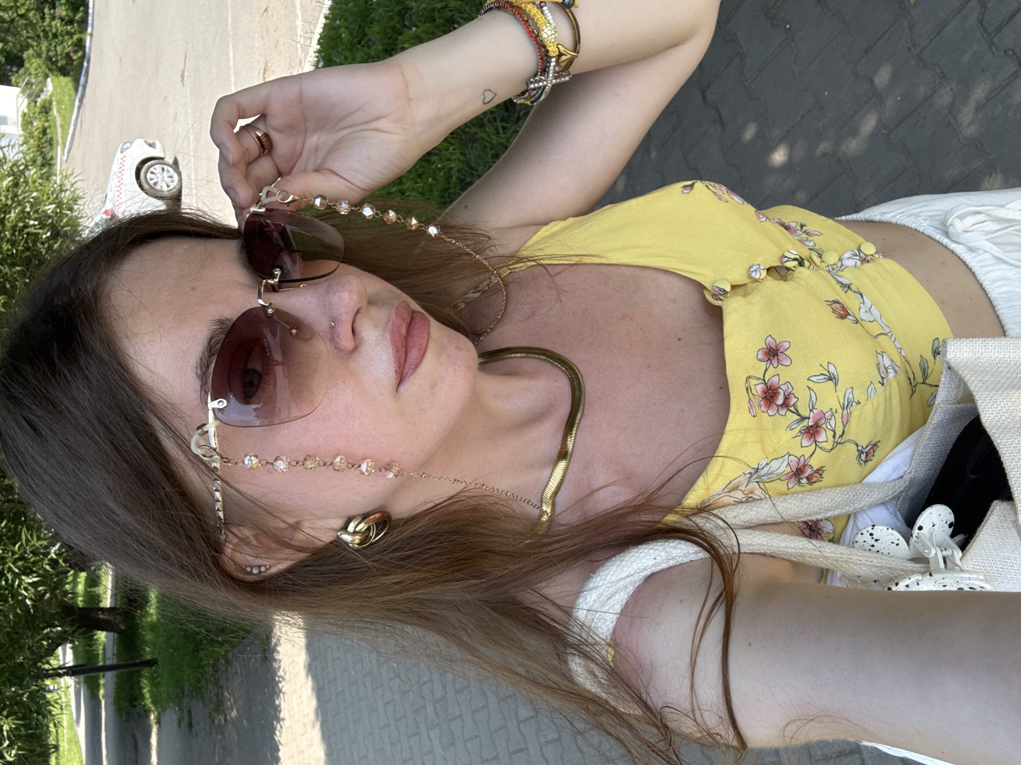 A woman with long brown hair is taking a selfie outdoors on a sunny day. She is wearing large, tinted sunglasses with a decorative chain made of beads and gold. She is also wearing large gold hoop earrings and a gold necklace. Her top is yellow with a floral pattern, and she has white pants. She is holding her sunglasses with her right hand and her left arm is visible. On her left arm, she has a tattoo of a small heart and several bracelets. She is carrying a canvas tote bag over her left shoulder. The background shows a paved walkway, grass, trees, and a white car with a red and white checkered pattern.
