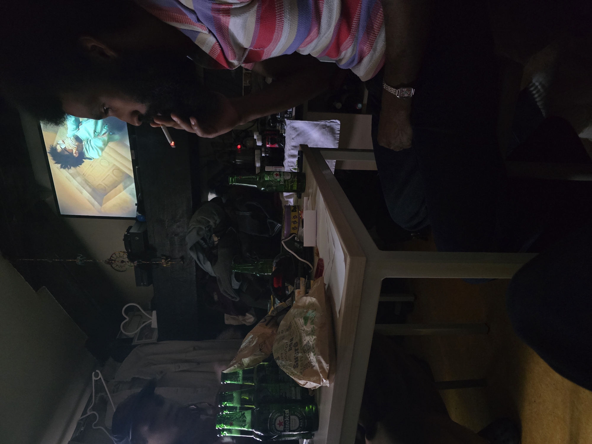 A person with dark hair and a beard is smoking a cigarette while watching a television screen that shows a man with an afro. The individual is wearing a striped shirt and a watch on their wrist. A white table is laden with empty green Heineken beer bottles and takeout bags. The environment is dimly lit, with clothing on hangers and a decorative heart visible in the background. The location appears to be indoors. There is no information to suggest this is in Paris, France.