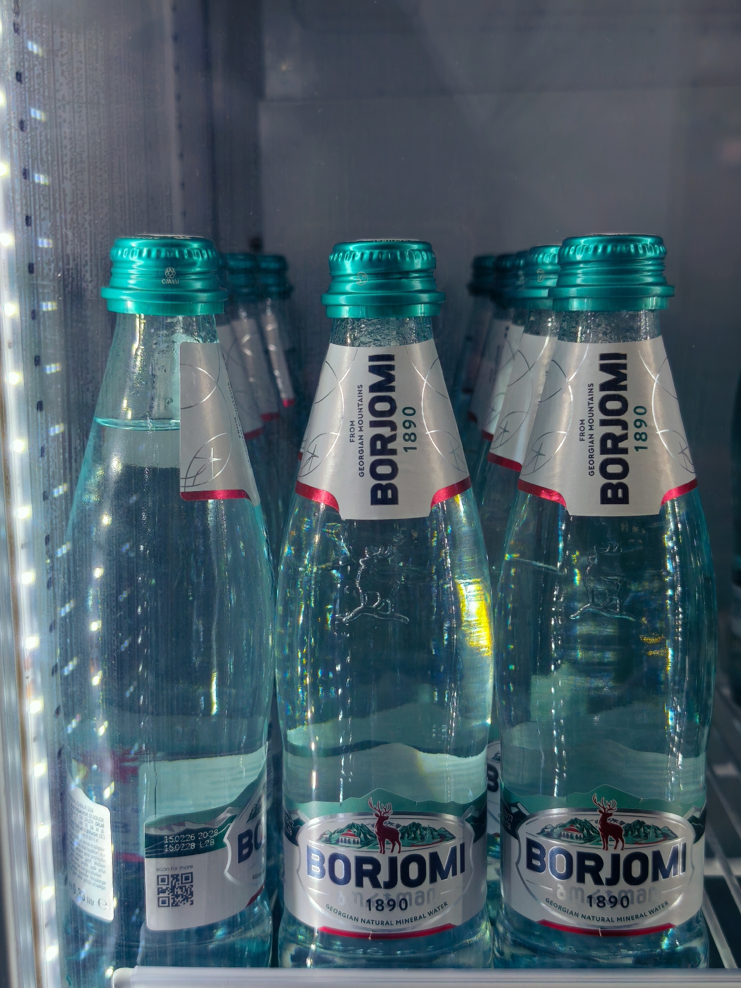 The image shows several bottles of Borjomi mineral water in a cooler with internal lighting. The bottles are made of clear blue-green glass and have distinctive teal-colored caps. Each bottle features a silver and white label with the Borjomi logo, the year 1890, and the text "FROM GEORGIAN MOUNTAINS" and "GEORGIAN NATURAL MINERAL WATER". Some bottles also display QR codes and expiry dates. The background appears to be a clean, refrigerated display unit, possibly in a store or convenience store. The lighting creates reflections and highlights on the bottles, indicating a bright and clean environment. The image is a close-up shot, focusing on the product. There are no people visible in the image, and no explicit details about the location in Tashkent, Uzbekistan are provided within the image itself, although it is implied to be a retail setting there.
