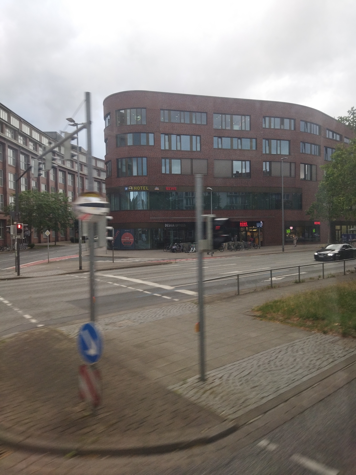 This is a medium-distance, eye-level, slightly blurred, horizontal, exterior, daytime photograph of a street scene in Hanover, Germany. The dominant subject is a large, curved, red brick building with numerous windows. The building has several businesses on the ground floor, including "REWE" (a supermarket chain) and "DESIGN OFFICES." A "P+P HOTEL" sign is also visible. To the left of the brick building is a taller, red brick building with a facade of many evenly spaced, light-colored windows. A traffic light with a red signal is visible on the left side of the frame. A blue and white traffic sign, indicating a mandatory right turn, is positioned on the sidewalk in the foreground. A black car is driving on the road in the middle ground. The sky is overcast and gray. The environment appears to be an urban commercial area with a street, sidewalk, and building facades.