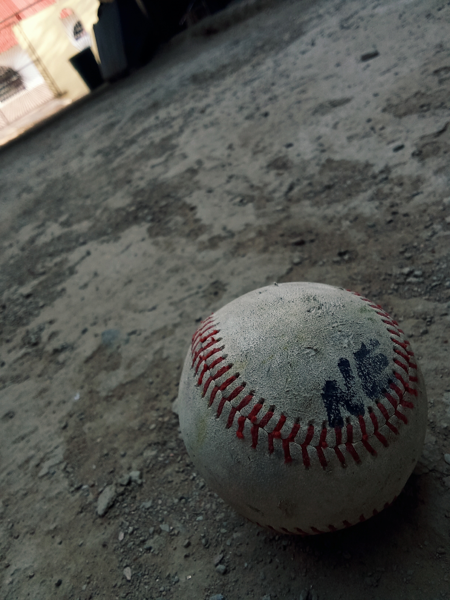 A well-worn baseball rests on a dirt and gravel surface. The ball is scuffed and marked with dark smudges. In the background, an indistinct structure is visible with a gated entrance. Given the prompt location of Juan Griego, Venezuela, the surroundings suggest a modest, possibly underdeveloped outdoor space in a residential area. There are no discernible people or actions evident. The scene is characterized by a discarded baseball in a somewhat desolate setting.