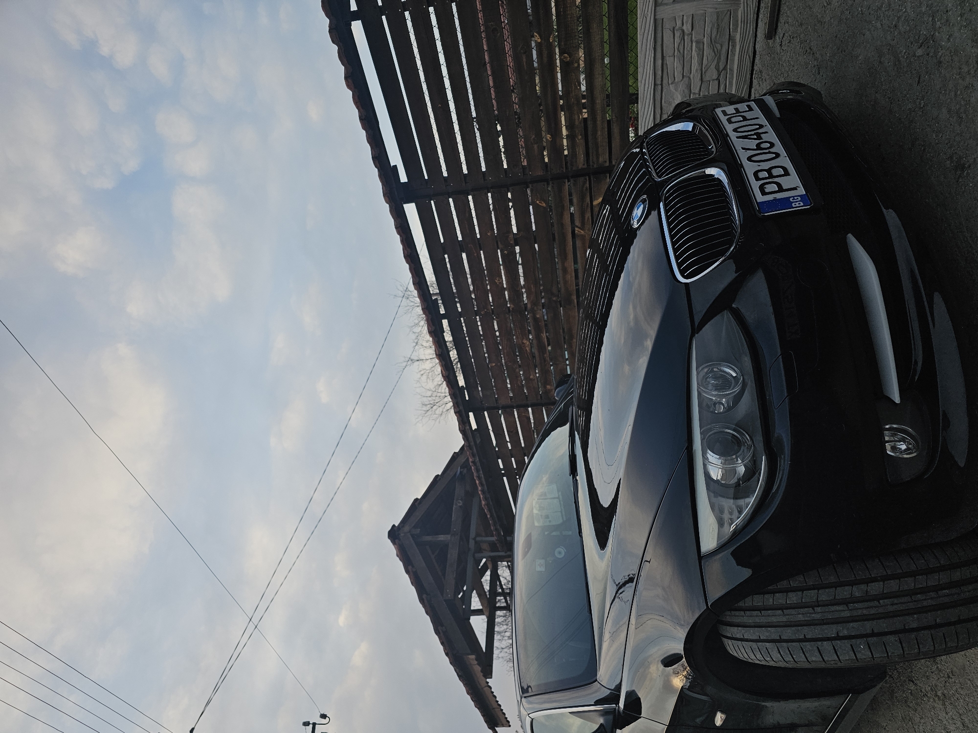 A black BMW car with a Bulgarian license plate (PB 0640 PE) is parked near a wooden structure in Krasnovo, Bulgaria. The sky above has a scattered cloud cover.