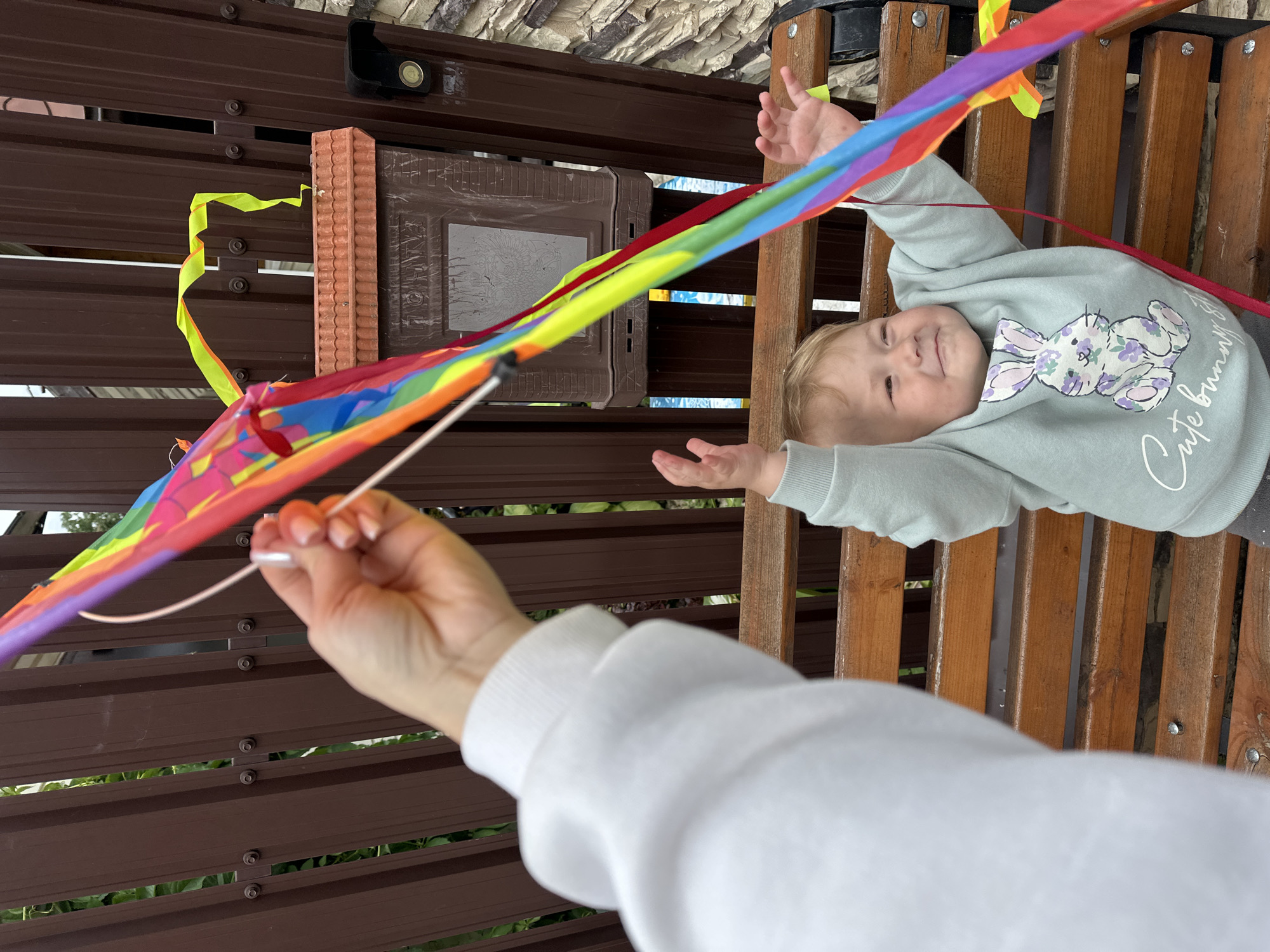 A person's hand, wearing a white sweatshirt, holds a string attached to a colorful kite, partially obscuring a young child sitting on a wooden bench. The child, wearing a light blue sweatshirt with a bunny design, looks up and to the side with arms raised. Behind them is a dark brown wooden fence and a brown mailbox. The scene appears to be outdoors, possibly in a residential area in Nizhny Novgorod, Russia.