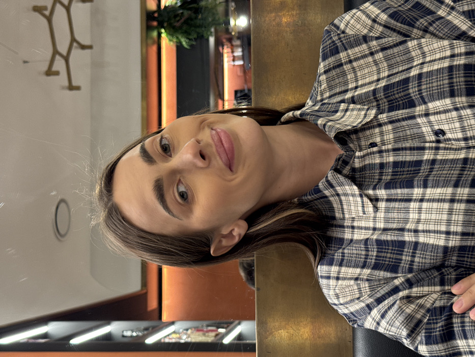 This image depicts a female subject indoors, situated in what appears to be a restaurant or bar in Verona, Italy. The subject, appearing to be in her 20s or 30s, is centered in the frame, looking directly forward with a subtle, closed-mouth smile. She has long, dark brown hair with lighter streaks and is wearing a blue and beige plaid collared button-up shirt. Her left arm is partially visible in the lower-left, suggesting she is seated. The background reflects various elements of an interior space on a polished or mirrored surface. To the left, a vertically illuminated shelving unit displaying bottles is visible. To the right, an orange-lit wall panel is present above a reflected bar counter featuring bottles and glassware. A large, gold-toned hexagonal light fixture is suspended from a white ceiling, which also shows a circular recessed light and reflected green foliage. A horizontal golden or brass-colored panel is positioned behind the subject's right shoulder.
