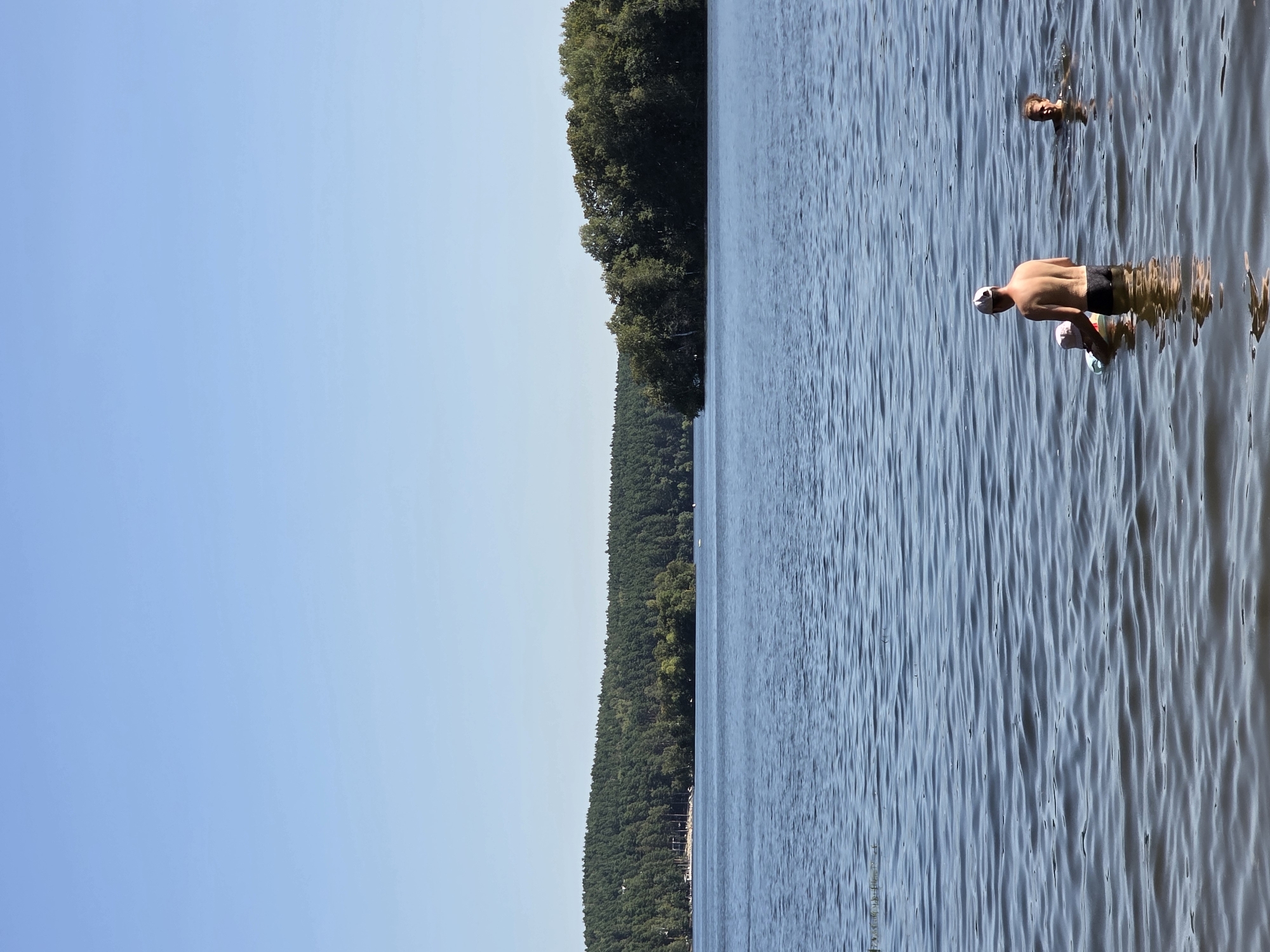 In the city of Chebarkul, Russia, on a bright, clear day, a serene lake dominates the scene. The water, a deep blue, reflects the cloudless sky and is gently rippled by a light breeze. On the far shore, a dense forest of green trees stretches across the horizon, interspersed with some lighter green deciduous trees, and a small, sandy beach with scattered structures and poles is visible to the left of the forest.
In the foreground, three people are enjoying the water. A man wearing a white baseball cap and dark swim trunks stands in the water, his back to the viewer. He appears to be supervising a young child wearing a pink hat and a floating aid. To the right of the man, another person, a woman, is swimming in the lake. Her head and shoulders are visible above the water. The overall impression is one of a peaceful summer day spent at a natural lake.