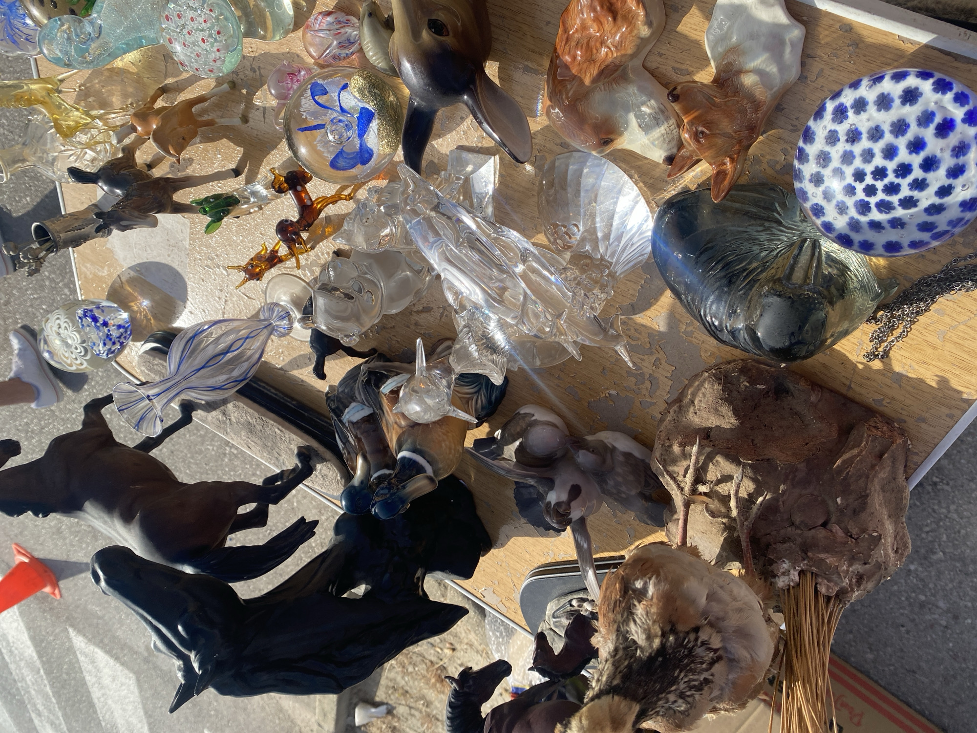 The image depicts an outdoor market stall, likely in Manilva, Spain, based on the sunlight and general atmosphere, though this is not explicitly confirmed by any signage or landmarks. The stall is laden with various decorative items, predominantly figurines and glass art. Several ceramic figurines are visible, including a group of ducks with detailed plumage, a pair of dog heads, and a fawn. There are also several glass objects, such as paperweights with intricate blue flower designs and abstract glass sculptures. A prominent black horse sculpture dominates the left side of the frame, with another brown horse figurine positioned behind it. In the foreground, a small bird figurine is perched on a natural, root-like base. The items are displayed on a wooden surface, which appears worn. Sunlight casts strong shadows, indicating it is daytime. No people are clearly visible in the immediate vicinity of the stall, though a pair of white sneakers is partially visible in the background, suggesting the presence of a shopper or vendor. The ground surface around the stall appears to be concrete or asphalt.