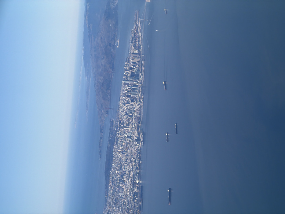 An aerial photograph captures a panoramic view of a major coastal city and its expansive bay under a clear, bright sky. The image, best viewed rotated clockwise by 90 degrees, places a densely developed urban area along the bottom and left side, stretching out into a vast body of water on the right.
The cityscape is characterized by numerous buildings, forming a skyline that indicates a bustling metropolitan center. In the mid-distance on the left, the iconic red-orange towers of the Golden Gate Bridge are visible, spanning a strait and connecting the main city to a hilly, mountainous landmass. This landmass, likely the Marin Headlands, is covered in natural terrain.
The wide body of water, indicative of a bay or estuary, is a deep blue and appears calm. Several large cargo ships or tankers are scattered across the water, either anchored or in transit, with a few smaller vessels also discernible by their faint wakes. The bright, even lighting suggests it is daytime, likely morning or afternoon, with excellent visibility. No people or legible text are visible due to the high altitude. The scene portrays a significant urban environment with substantial maritime activity, strongly identifying the location as San Francisco Bay, consistent with the provided Oakland context.