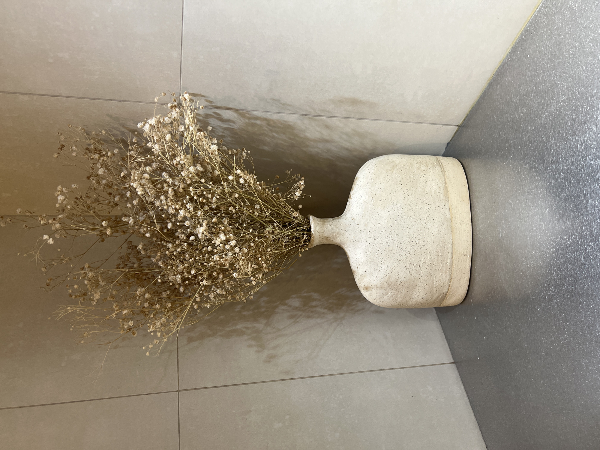A dry floral arrangement is placed in a white, ceramic vase with a narrow neck and bulbous body. The vase is resting on a tiled floor, a light-colored tan in one area transitioning into a gray tile closer to the wall. The floor tiles are rectangular. The origin of the image is Yerevan, Armenia.