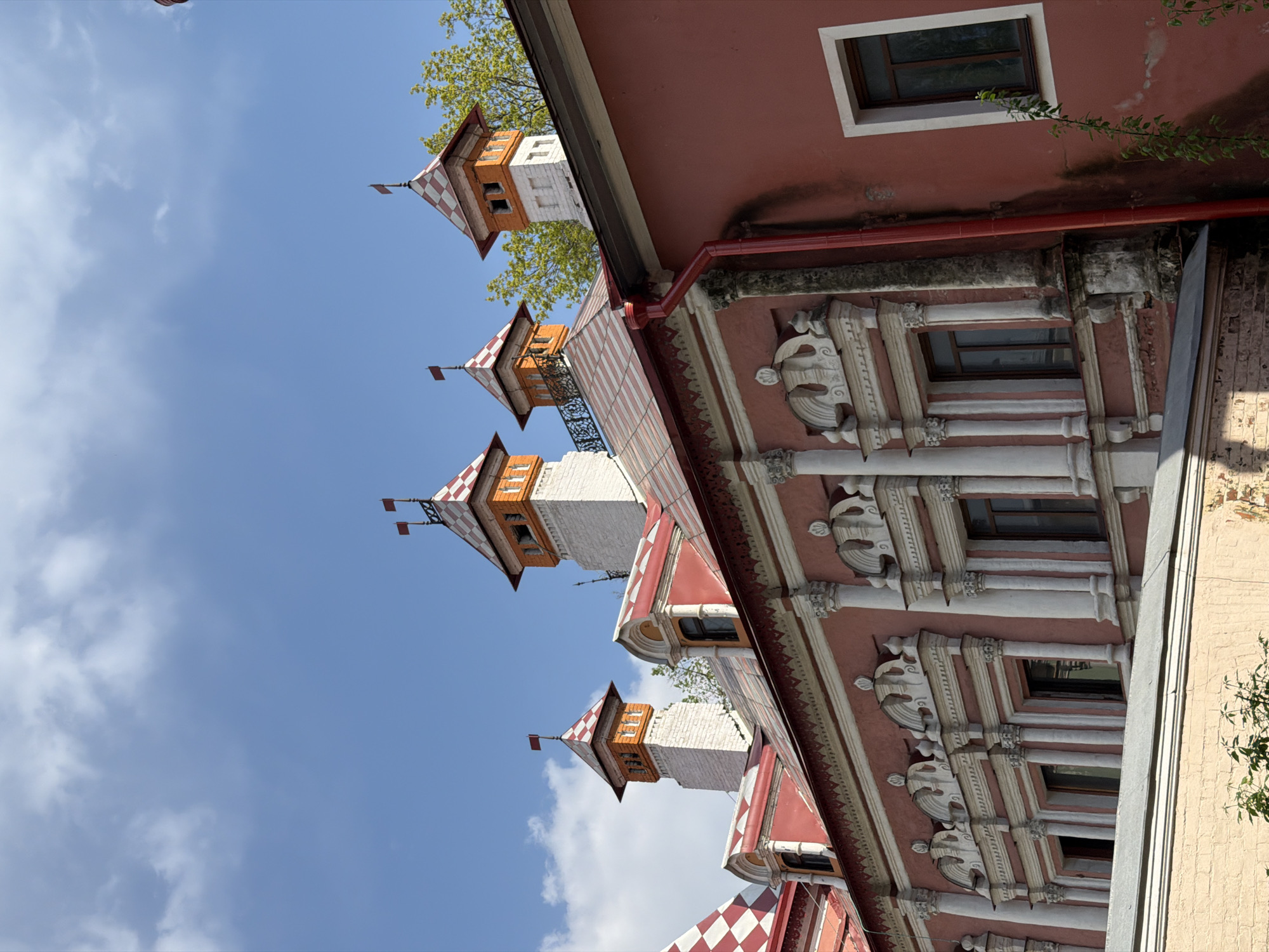 This image depicts the exterior of a building with distinct architectural features. The building has multiple decorative turrets, each topped with a red and white checkered roof and a small flag. The towers are primarily white with orange accents.
The building's facade is predominantly red with white decorative elements, including columns and sculpted details around the windows. There are several windows visible, some with visible reflections of the interior.
The background includes a blue sky with scattered white clouds and some glimpses of green foliage, possibly from nearby trees. The perspective of the image is from a low angle, looking upwards toward the building.
The building is located in Moscow, Russia.