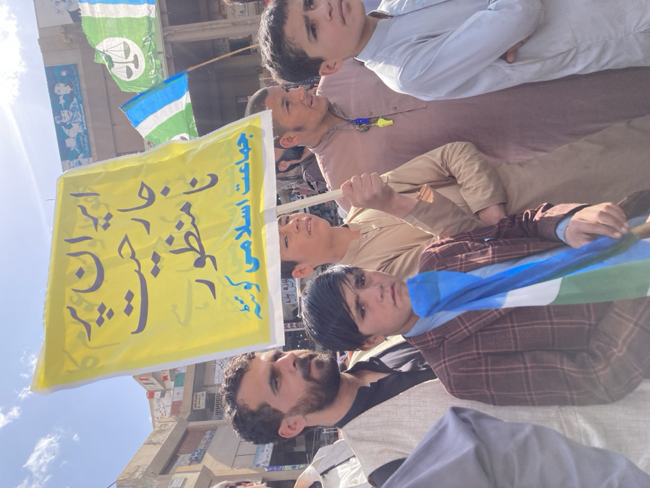 The image depicts a daytime outdoor gathering of approximately six males in the foreground and numerous others in the background, situated in Quetta City Tehsil, Pakistan. The scene is illuminated by bright daylight, with a clear blue sky and some white clouds overhead.
In the left foreground, a man with a dark beard and mustache, wearing a black collared shirt under a light-colored waistcoat, looks towards the right. To his right, a young boy with dark hair, dressed in a brown plaid blazer over a light blue collared shirt, holds a folded blue, white, and green striped flag-like fabric in his left hand and gazes directly forward.
Centrally, a teenage boy in a light brown shalwar kameez holds a large, rectangular yellow sign vertically with both hands. The sign features Urdu text in dark blue, which reads from top to bottom: "ایران پر جارحیت نامنظور" (Aggression on Iran Unacceptable/Rejected) and "جماعت اسلامی کوئٹہ" (Jamaat-e-Islami Quetta). This boy looks towards the right.
To the right of the boy holding the yellow sign, a man in a dark brown/mauve shalwar kameez wears a yellow whistle on a lanyard around his neck and looks rightward. Further to the right in the foreground, another young boy in a light grey shalwar kameez stands with his arms crossed and looks towards the right.
In the background, multi-story urban buildings are visible, some appearing weathered. On a prominent building facade, a large banner or poster featuring images of individuals is discernible. To the right of the yellow sign, multiple green flags with a white scale emblem, indicative of the Jamaat-e-Islami party, and other blue and white striped flags are present, held by unseen individuals within the larger crowd. The gathering appears to be a protest or rally organized by Jamaat-e-Islami.