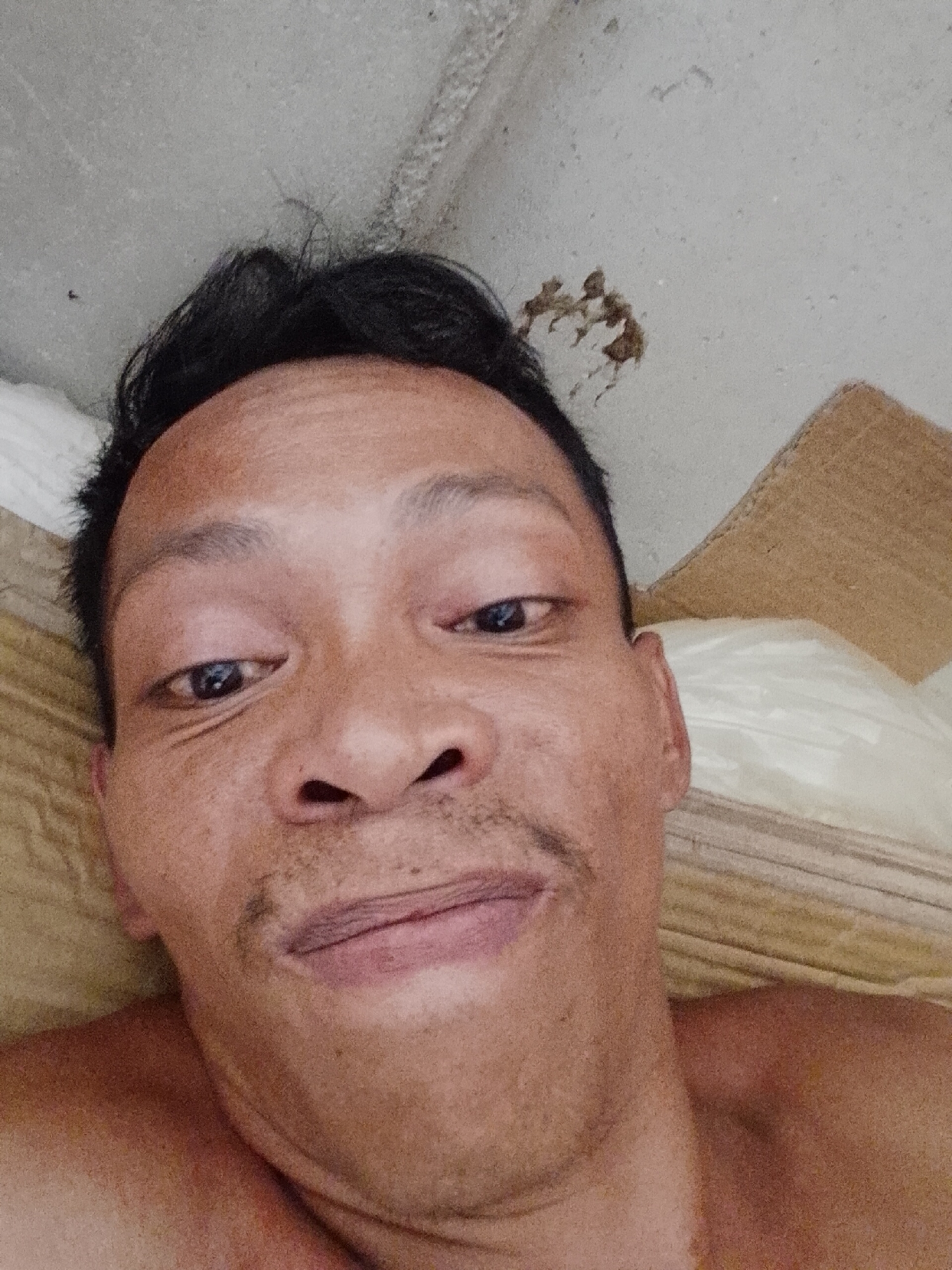 A close-up, selfie-style image shows a middle-aged Asian man with dark hair, a slight mustache, and a serious expression. He appears to be lying down on a bed with patterned tan and white sheets and a tan pillow visible behind him. The image is taken from a low angle, with the ceiling partially visible in the upper left corner. There are what appears to be dark splatters on the ceiling. The man's shoulders and upper chest are visible, suggesting he is shirtless. There are no indications of the specific location in Bukit Ketok, Indonesia.