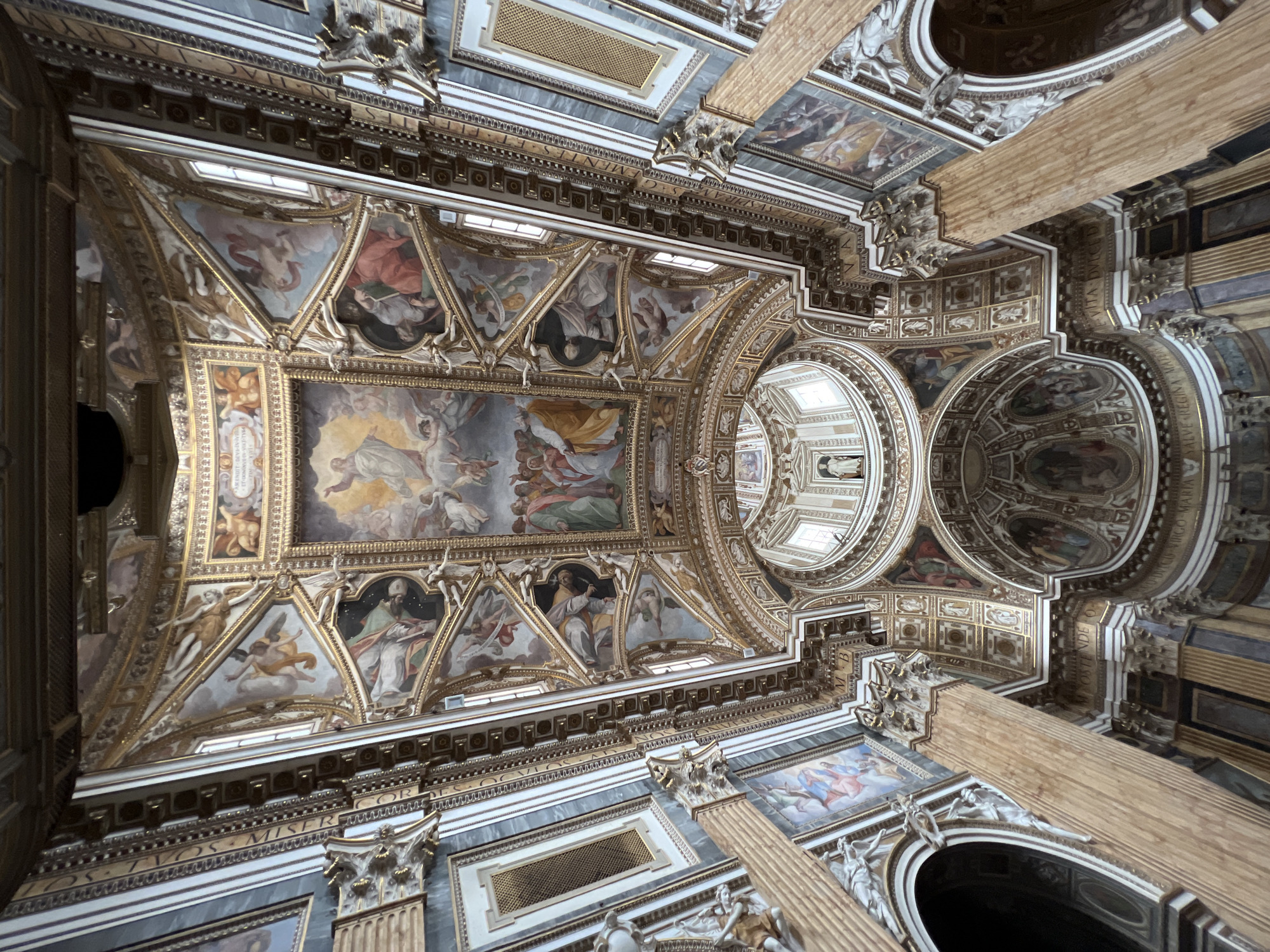 This media file captures an awe-inspiring, upward-looking view of the highly ornate vaulted ceiling and upper architecture of a grand church or basilica, a style characteristic of structures found in Rome, Italy.
**Subjects:** The predominant subjects are the numerous fresco paintings that adorn the ceiling. These artworks depict various religious scenes and figures, including angels, often as putti, saints, and divine figures in celestial settings with clouds and rays of light, suggesting ascension or heavenly glory. The paintings are meticulously framed within richly gilded, coffered sections and arches. Surrounding these artworks are intricate architectural details: decorative moldings, cornices, pilasters, and large, fluted columns or pilasters that rise towards the ceiling. A large central dome, featuring additional painted sections and smaller windows, is visible towards the right, along with other smaller domes or apses.
**Activity or event taking place:** The frescos portray religious narratives, likely depicting episodes from the lives of saints or biblical events, emphasizing themes of divine intervention, salvation, and heavenly hosts. The dynamic poses of the painted figures suggest movement and spiritual elevation.
**Notable details:** The entire structure is characterized by overwhelming opulence, with extensive use of gold leaf on frames, friezes, and decorative elements. This opulent gold contrasts strikingly with the lighter tones of the painted surfaces and the grey and white marble-like architectural components. Natural light filters in through high-set windows, illuminating the intricate details and enhancing the gilded surfaces, creating a sense of immense height and grandeur.
**Visible text:** Several Latin inscriptions are discernible along the friezes and cornices. Portions of the prayer "SALVE REGINA MATER MISERICORDIAE" (Hail, Holy Queen, Mother of Mercy) can be seen, along with other fragments such as "...ORUM TUOS MISER..." and "...OSTE... M VNDI...", which further affirm the religious context and possibly hint at specific dedications or theological messages.