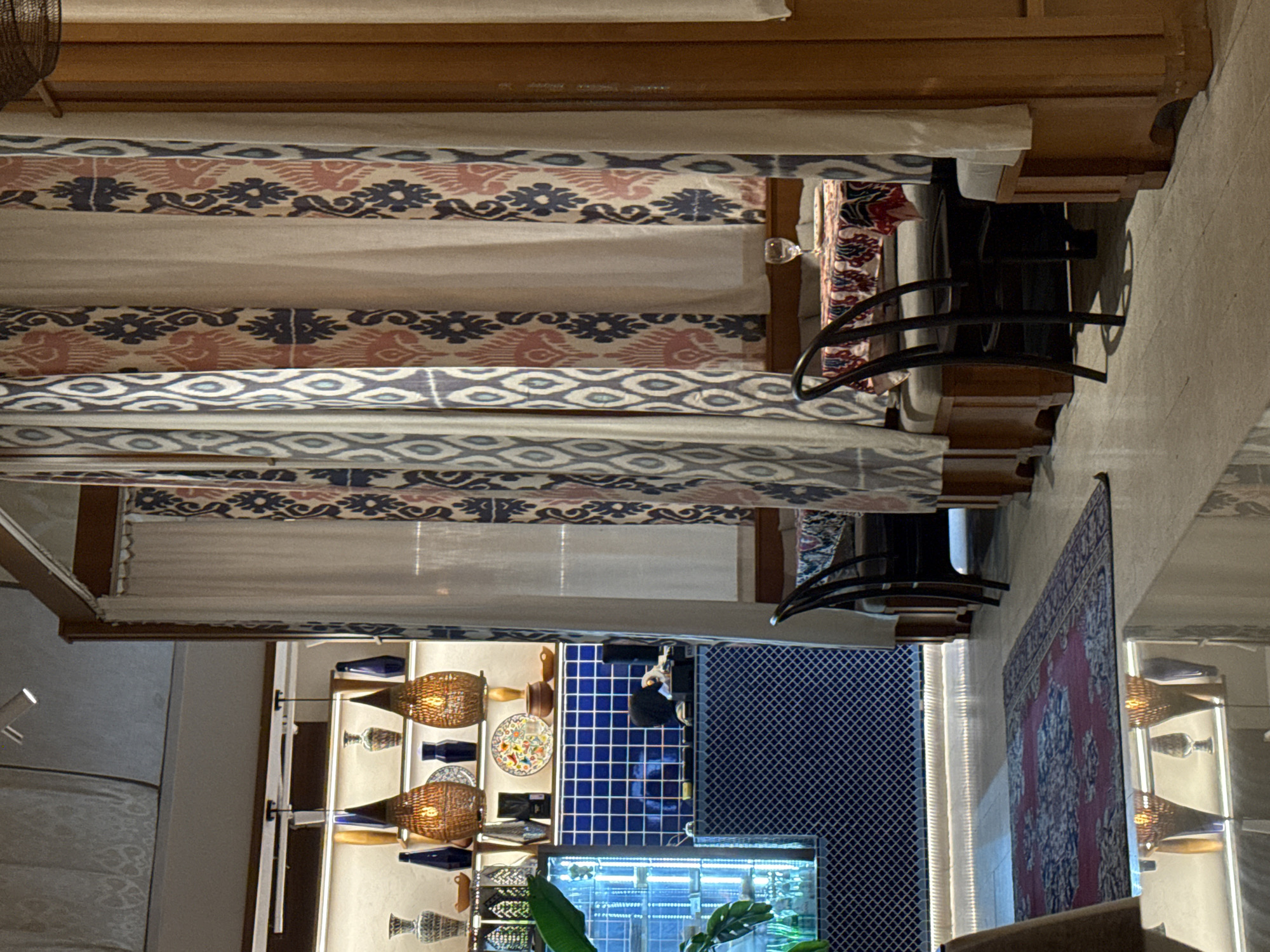 This media file captures an elaborately decorated indoor space, likely a restaurant, lounge, or a section of a hotel, characteristic of high-end interiors often found in Dubai. The scene is brightly lit by various artificial light sources, suggesting it was taken during the evening or night.
The foreground is dominated by a series of curtains featuring traditional Ikat-style patterns with stylized floral and geometric motifs in shades of blue, dusty pink, and cream, hanging from a sturdy wooden frame, possibly a canopy or divider. To the right, a dark, elegant chair with curved armrests is positioned next to a small, dark table holding a clear glass. The floor appears to be a light, neutral-toned surface.
Towards the mid-ground and background, the room opens up to reveal more decorative elements. On the left, several woven pendant lights, emitting a warm, soft glow, are mounted alongside shelves displaying various decorative items, including deep blue bottles or vases and colorful patterned plates. This area transitions into a striking feature wall covered in small, vibrant blue square tiles. Below this, an illuminated display cabinet or counter showcases additional decorative objects and foliage, with lush green plant leaves visible.
On the right side of the image, a large, intricately patterned rug, predominantly red with blue and cream floral designs, is prominently displayed, hanging on a wall. Below it, another illuminated display unit is visible, featuring more woven items and decorative vases, further enhancing the rich, curated aesthetic of the space.
No people are present in the image, implying a moment of quiet or preparation within the setting. There is no visible text. The sophisticated blend of traditional patterns, warm lighting, and decorative accents creates an inviting and culturally rich ambiance.