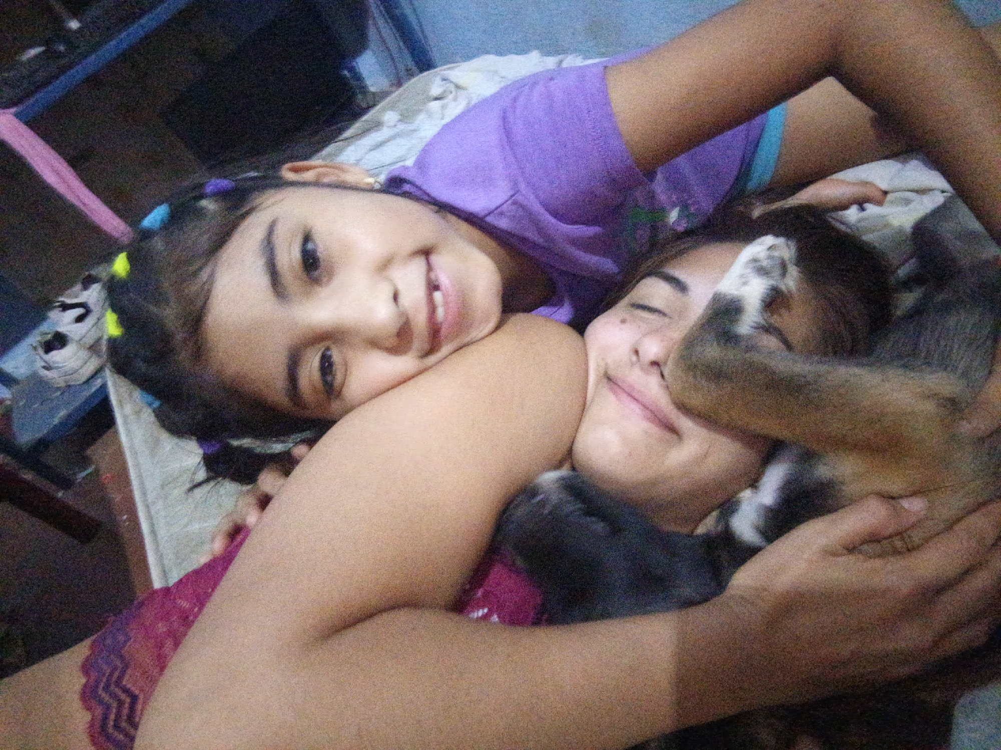 Two females are lying on a bed indoors. The younger female is wearing a purple shirt and has colored hair ties in her hair, and is smiling at the camera. The other female has her eyes closed and appears to be holding a puppy or dog. The location is Juan Griego, Venezuela.