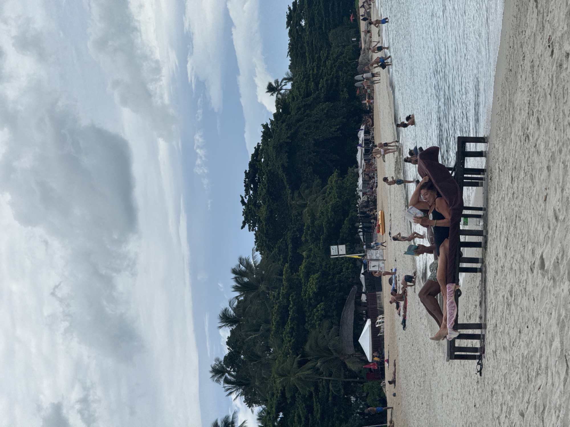 The image depicts a beach scene in the Ko Samui District, Thailand. In the foreground, two individuals are relaxing on loungers on the sand. One person is lying on their back with their head propped up on a cushion, holding a book or phone. The other person is lying on their stomach. Behind them, a shallow body of water is visible, with several people swimming and wading. Further back, the sandy beach is populated with more individuals, some sitting under umbrellas, and others walking. A line of lush green trees and palm trees forms a dense backdrop along the coast. In the distance, there are some small structures that appear to be huts or businesses. The sky is overcast with scattered clouds.