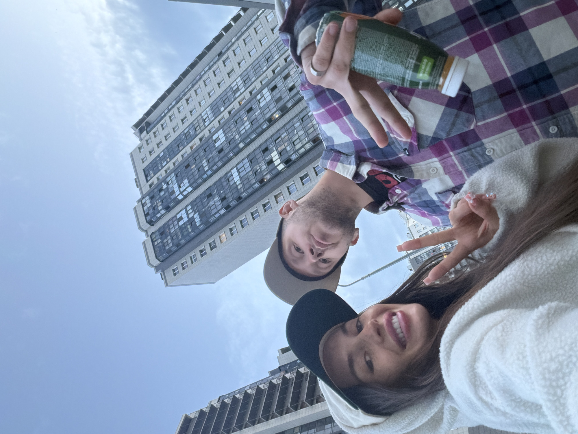 In the foreground, a woman wearing a white hooded jacket and a baseball cap smiles widely, sticking out her tongue slightly. She is making a peace sign with her right hand. Beside her, a man wearing a plaid shirt and a baseball cap poses, holding a green bottle and making a peace sign with his left hand. They are both looking up and slightly towards the camera, with tall apartment buildings in the background. The sky is overcast.