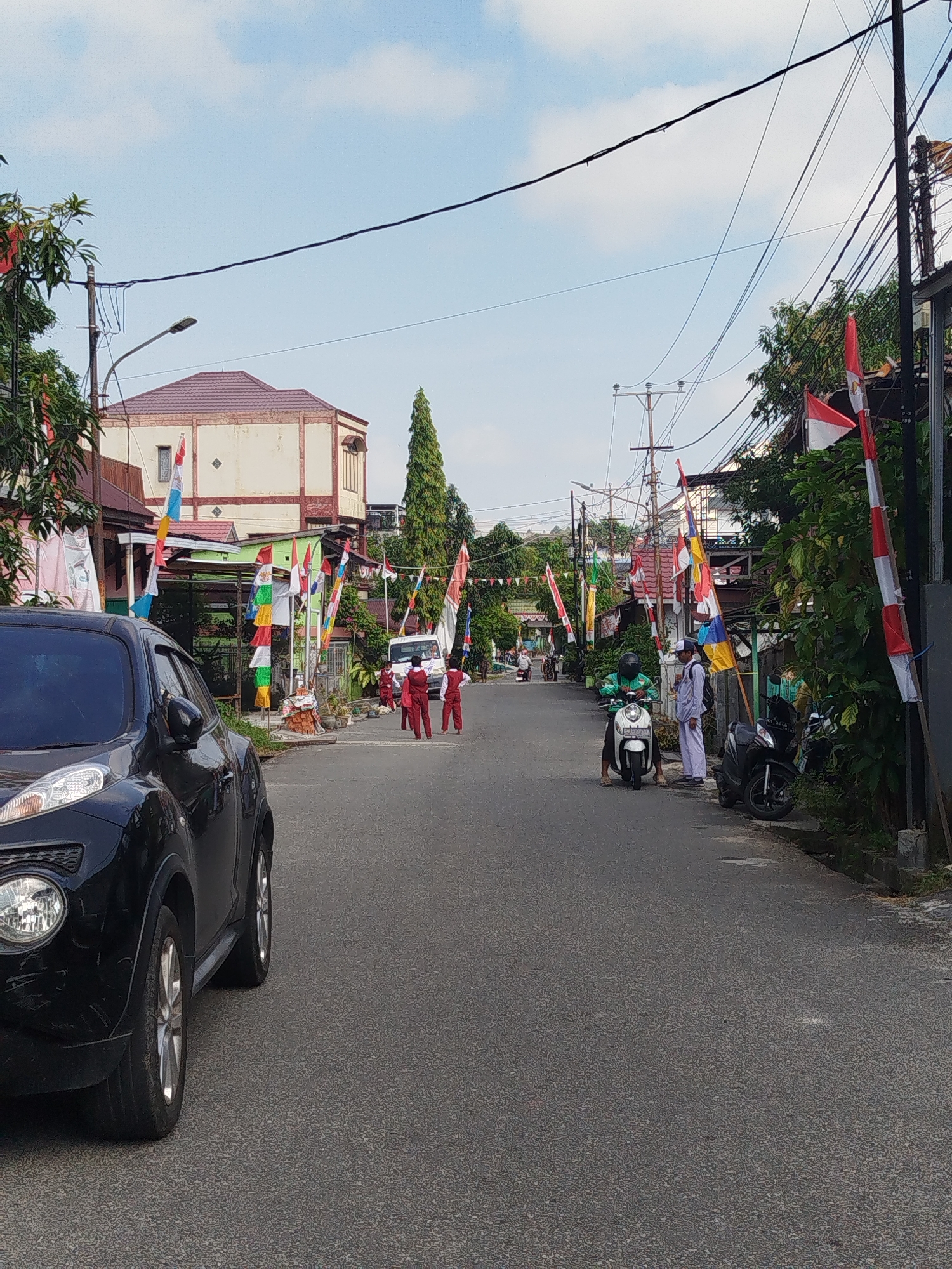 The image shows a residential street in Balikpapan, Indonesia, decorated with numerous red and white flags, commonly seen during national celebrations like Independence Day. The street is paved and slopes slightly uphill. On the left side of the street, a black car is parked, partially visible. Further down the street, several people are gathered, some in red and white uniforms, possibly students. There are also several motorcycles parked along the street. Buildings line both sides of the street, with some featuring red tile roofs. Overhead, electrical wires are strung between poles, common in urban areas. The sky is blue with some clouds.