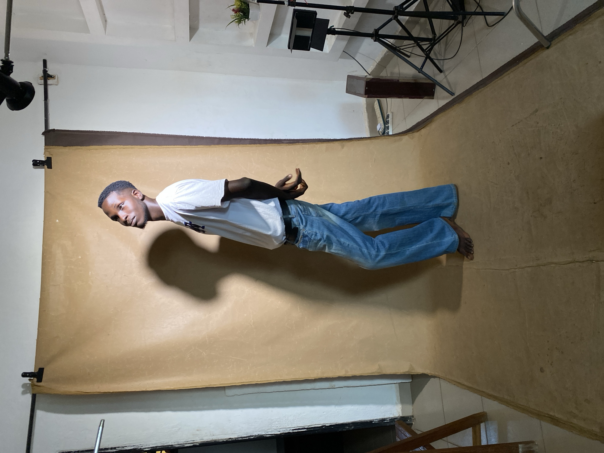 A young Black man is depicted standing in a photo studio in Bukuru, Nigeria. He has short dark hair and a light beard, and is looking towards the left of the frame with a neutral expression. He is wearing a white short-sleeved t-shirt, blue jeans, and is barefoot. His hands are clasped in front of his waist in a relaxed pose.
The man is positioned on a light brown or beige seamless paper backdrop that extends from the wall to the floor, showing some creases and wear. The studio setting is evident from the visible equipment. To the left, a black light stand with an attached light fixture is seen. To the right and upper background, another black tripod-style light stand supports a rectangular light fixture, with a small potted plant visible behind it. A dark wooden bench or box is also present on the right side. The ceiling above is white with recessed panels, and the floor beyond the backdrop is light-colored tile. The man's shadow is cast prominently on the backdrop behind him, indicating studio lighting. There is no visible text.