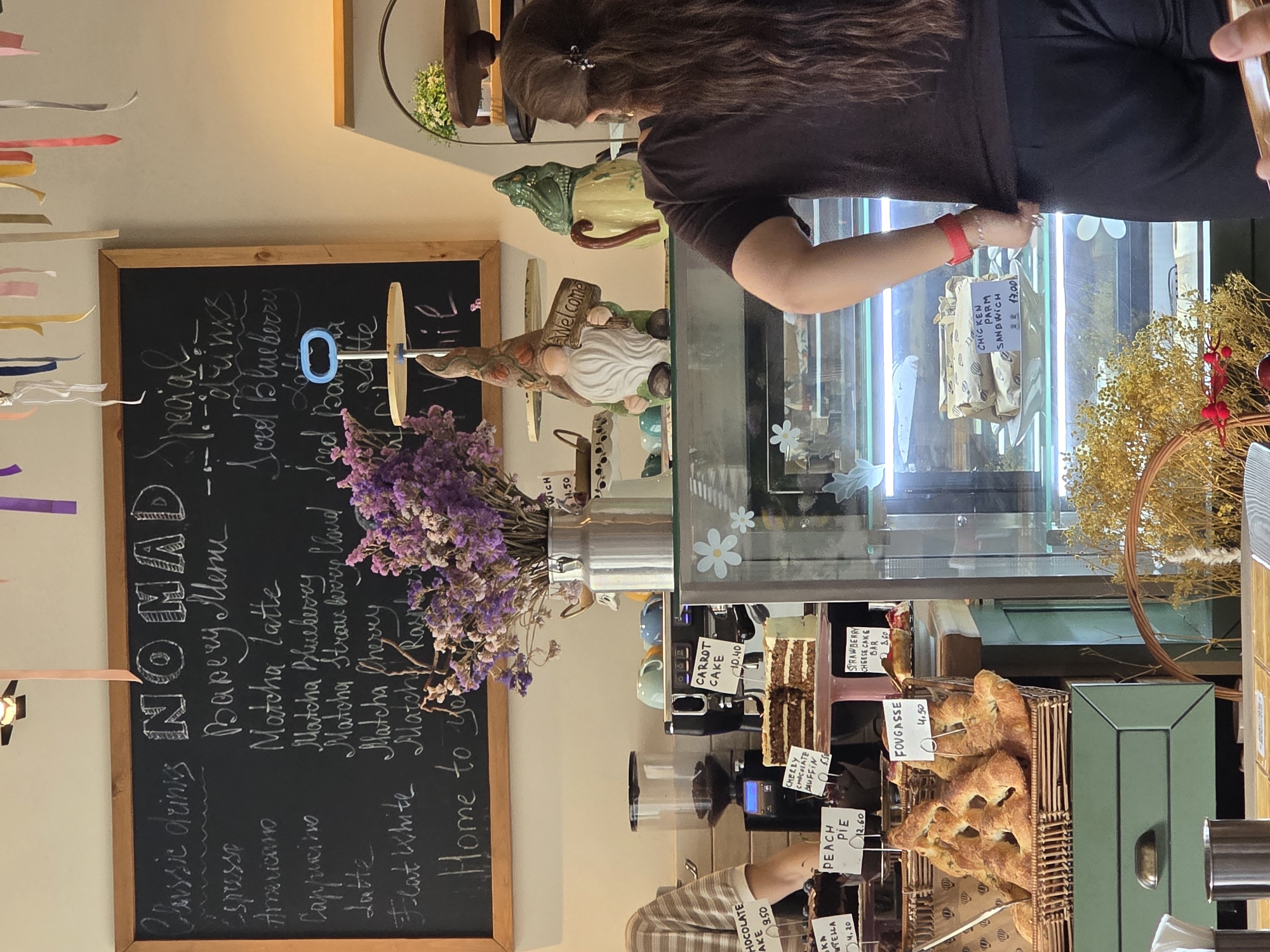 The image depicts the interior of a cafe, likely in Khatay Raion, Azerbaijan, judging by the presence of items commonly found in such establishments. A blackboard menu is prominently displayed, listing "Classic drinks" and "Bakery Menu." Items like espresso, americano, cappuccino, latte, and flat white are noted under classic drinks, while the bakery section features matcha latte variations, iced blueberry latte, iced banana latte, and a dessert named "Fougasse" with a price of 4.50.
The cafe appears to be decorated with whimsical elements. A gnome figurine holding a "Welcome" sign stands near a frog-shaped ceramic pitcher, adding a playful touch to the counter area. In the foreground, a display case showcases various baked goods, including a large loaf of Fougasse and slices of cake. One cake, labeled "CARROT CAKE," is priced at 10.40, and another, a "STRAWBERRY CHEESE CAKE BAR," is 8.60. Further inside the display case, pre-packaged sandwiches, including a "CHICKEN PARM SANDWICH" at 17.00, are visible.
A person, seen from behind and partially to the left, is interacting with the cafe's offerings or counter. They are wearing a dark top and dark trousers, with their long dark hair tied back with a clip. A red wristband is visible on their left wrist. The cafe environment seems cozy, with a mix of modern equipment, like a coffee grinder and espresso machine, and rustic decor, such as dried flowers in a milk jug vase. The overall lighting suggests a daytime scene, with warm ambient light enhancing the atmosphere. The presence of a metal trash can and a wooden table in the foreground suggests this is a functional space for customers.