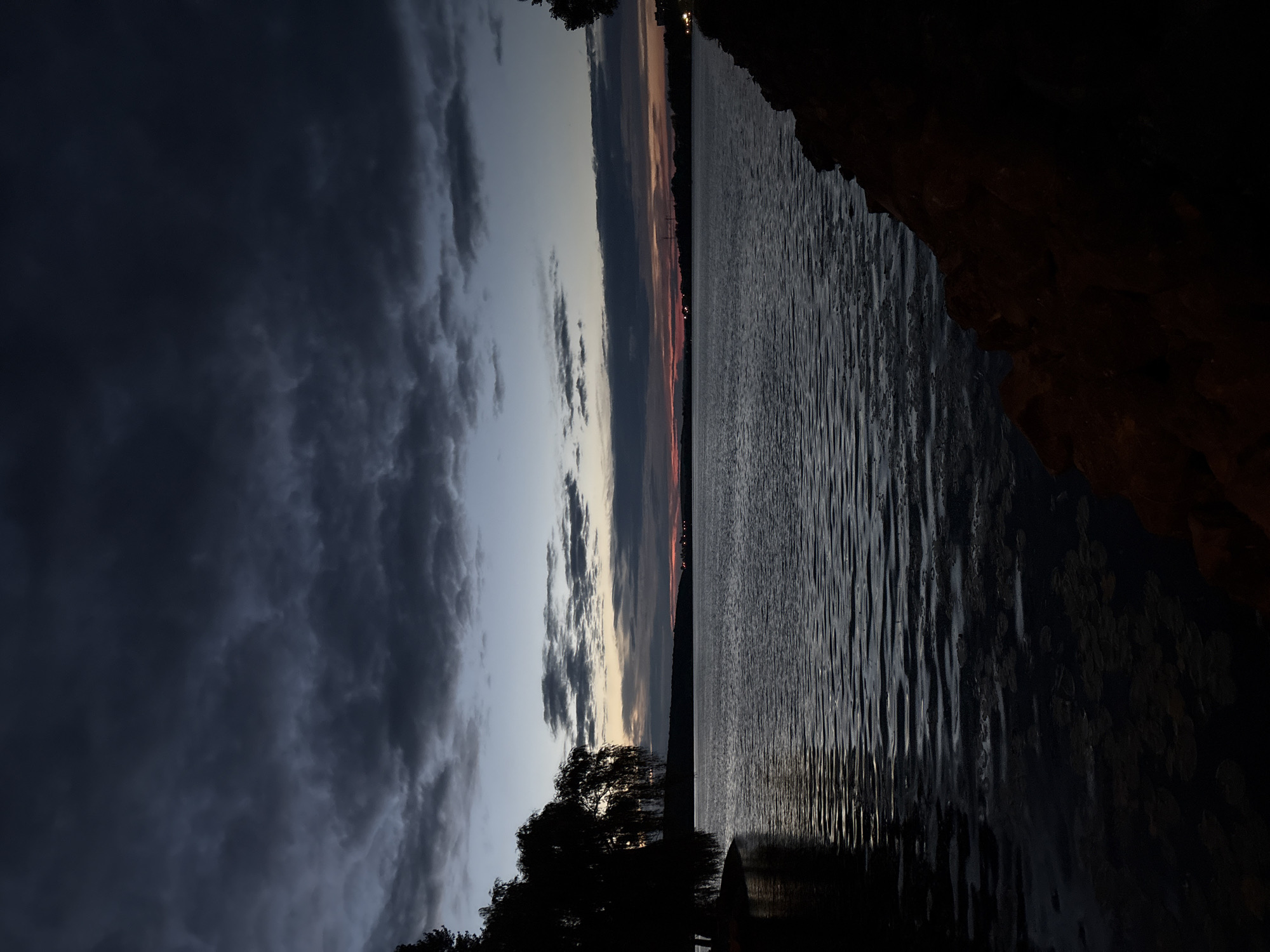 The image depicts a twilight scene over a body of water, likely a lake or river, bordering a city. The sky is dominated by dark, heavy clouds, with a sliver of pale light visible on the horizon, tinged with faint reds and oranges, suggesting the last moments of sunset or the early signs of dawn. The water surface is rippled, reflecting the dim sky and casting dark shadows. On the left side of the frame, the silhouette of a large, weeping willow tree is prominent, its branches drooping towards the water. The right side of the image features a dark, rocky shoreline. In the distance, across the water, faint lights of a city are discernible, indicating a populated area. The overall atmosphere is somber and serene. This scene is located on the Ternopil Reservoir, on the Seret River, adjacent to the city of Ternopil, Ukraine.