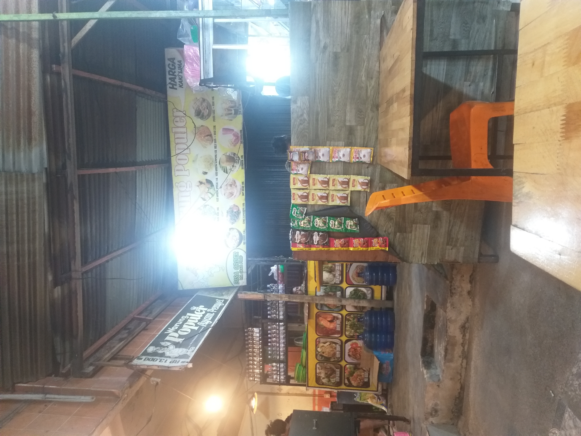 The image depicts an outdoor food stall at night. The stall is named "Warung Populer Ayam Penyet" and features a menu board with images of various dishes and their prices. Several rows of bottled drinks are displayed on shelves behind the counter. In front of the stall, there are large blue water dispensers and a yellow kiosk displaying food items. Packets of snacks and drinks are arranged on a wall to the right of the stall. An orange plastic chair is placed next to a wooden table, suggesting a seating area for customers. The overall ambiance is that of a casual, street-side eatery. Based on the street signs and the name of the establishment, it is likely located in Dumai City, Indonesia.