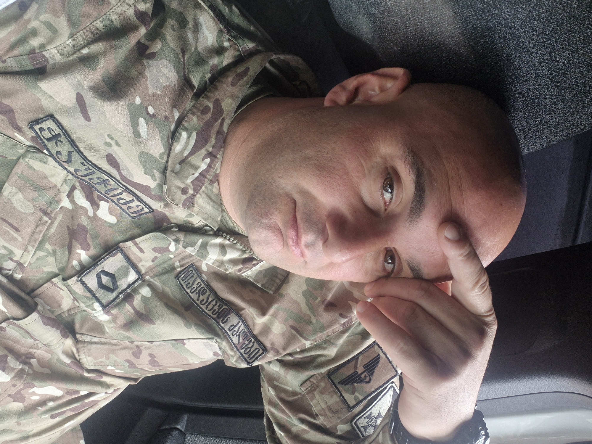 A close-up, slightly angled portrait of a bald man in military camouflage fatigues. The camouflage pattern is a blend of greens, browns, and tans. On the left breast of his uniform is a name tag with text in the Georgian alphabet and the Latin alphabet: “S-5 TDPPODE.” Another patch on the left breast features a gray diamond symbol. Another patch has text in the Georgian alphabet that appears to indicate a location. Below it is a patch with a winged design. He wears a black watch. His head is slightly tilted to the left, and his fingers touch the right side of his forehead. The background appears to be dark gray upholstery. The location is not explicitly visible, so there is no verifiable information that would definitively place the location in the city of Tbilisi, Georgia.