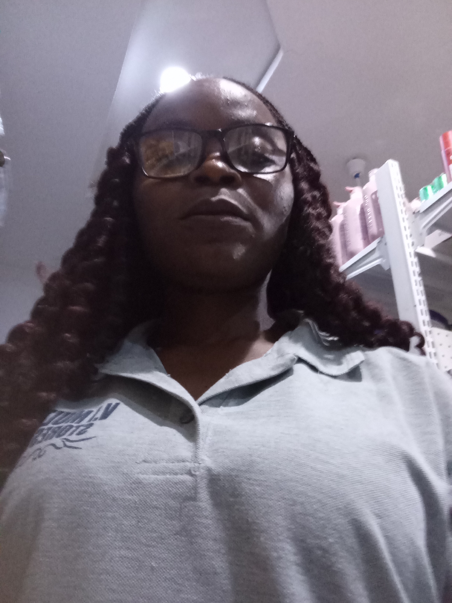 The media file is a first-person, slightly blurry selfie of a Black woman wearing glasses and a grey collared shirt. She has long, dark, braided hair that falls around her shoulders. The background appears to be an indoor environment, possibly a store or a warehouse, with shelves stocked with various products visible in the upper right portion of the image. The lighting suggests it could be daytime, but the indoor setting makes it difficult to determine. The woman's expression is neutral. On her shirt, there is some visible text in a dark blue font, which appears to be a logo or branding, but it is partially obscured and difficult to read entirely. The text seems to include the words "GLOBAL" and "SPORTS" along with numbers, possibly "21085" and "23012". The overall scene is casual and appears to be a moment captured in a retail or storage setting.