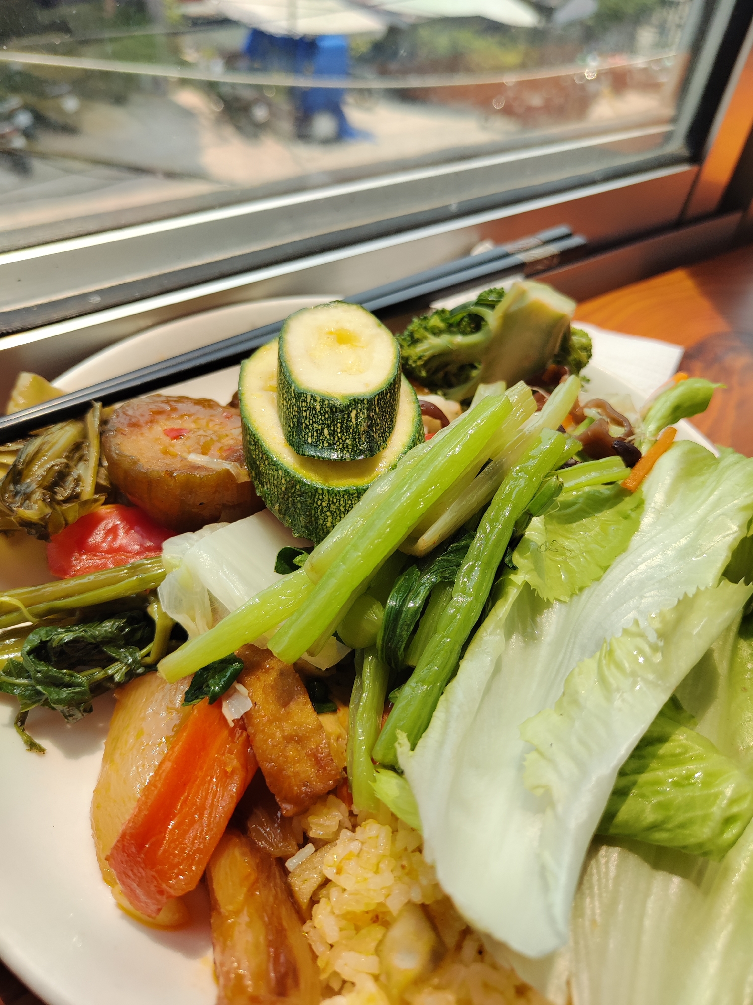 A plate of food is positioned on a brown wooden surface, likely a table, with a window behind it. The food includes cooked rice, various stir-fried and raw vegetables, zucchini slices stacked on top of each other, broccoli, and what appears to be tofu and pickled vegetables. Chopsticks are resting on the edge of the plate. The lighting suggests an indoor setting, with natural light filtering through the window. Outside the window, one can glimpse elements of an urban setting. Based on the provided information, the location is Da Lat, Vietnam.