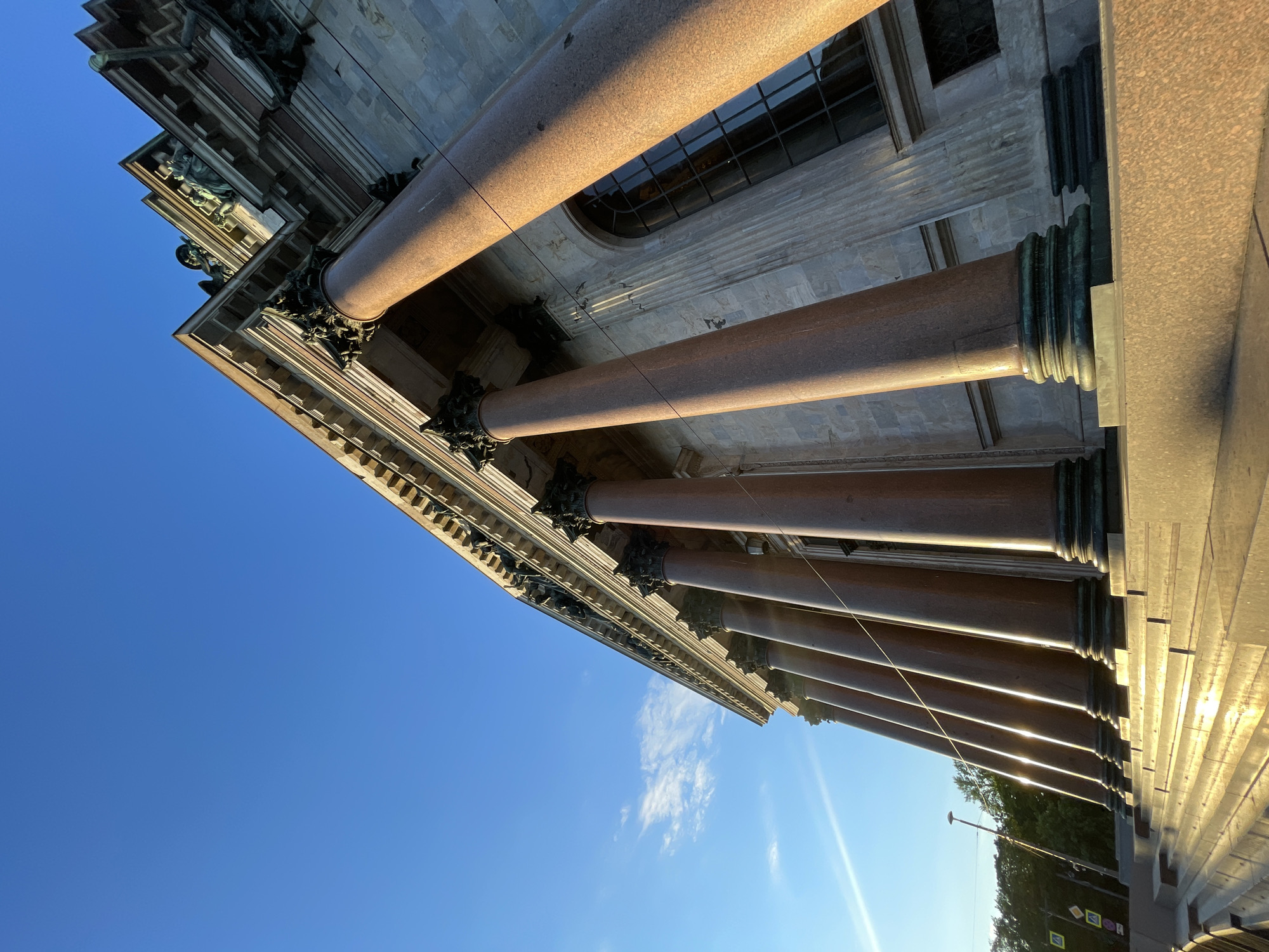 This low-angle, wide-shot photograph captures the imposing facade of St. Isaac's Cathedral in Saint Petersburg, Russia, bathed in the warm glow of early morning sunlight. The sky is a brilliant, cloudless blue, with a faint contrail visible high above. A single, wispy cloud drifts near the horizon on the left side of the image.
Dominating the frame are the massive, polished red granite columns that support the cathedral's portico. These columns, detailed with dark green bronze bases and intricately carved capitals, stretch upwards towards the sky. The sun's light catches the polished surfaces of the columns, creating strong highlights and casting long shadows.
The cathedral's facade is constructed of stone blocks, with classical architectural details visible along the top. Sculptures, seemingly figures, are perched on the cornices. A large, arched window with a dark grid pattern is visible behind the columns.
In the foreground, a wide staircase made of granite leads up to the cathedral. The steps are also illuminated by the sun, with the light reflecting off their surfaces.
To the left of the cathedral, a line of trees forms a dark green backdrop. A modern lamppost stands tall among the trees, and two yellow traffic signs with black symbols are mounted on poles, indicating pedestrian crossings.
The overall impression is one of grandeur, architectural splendor, and a serene morning in the historic city. There are no people or animals visible in the photograph.