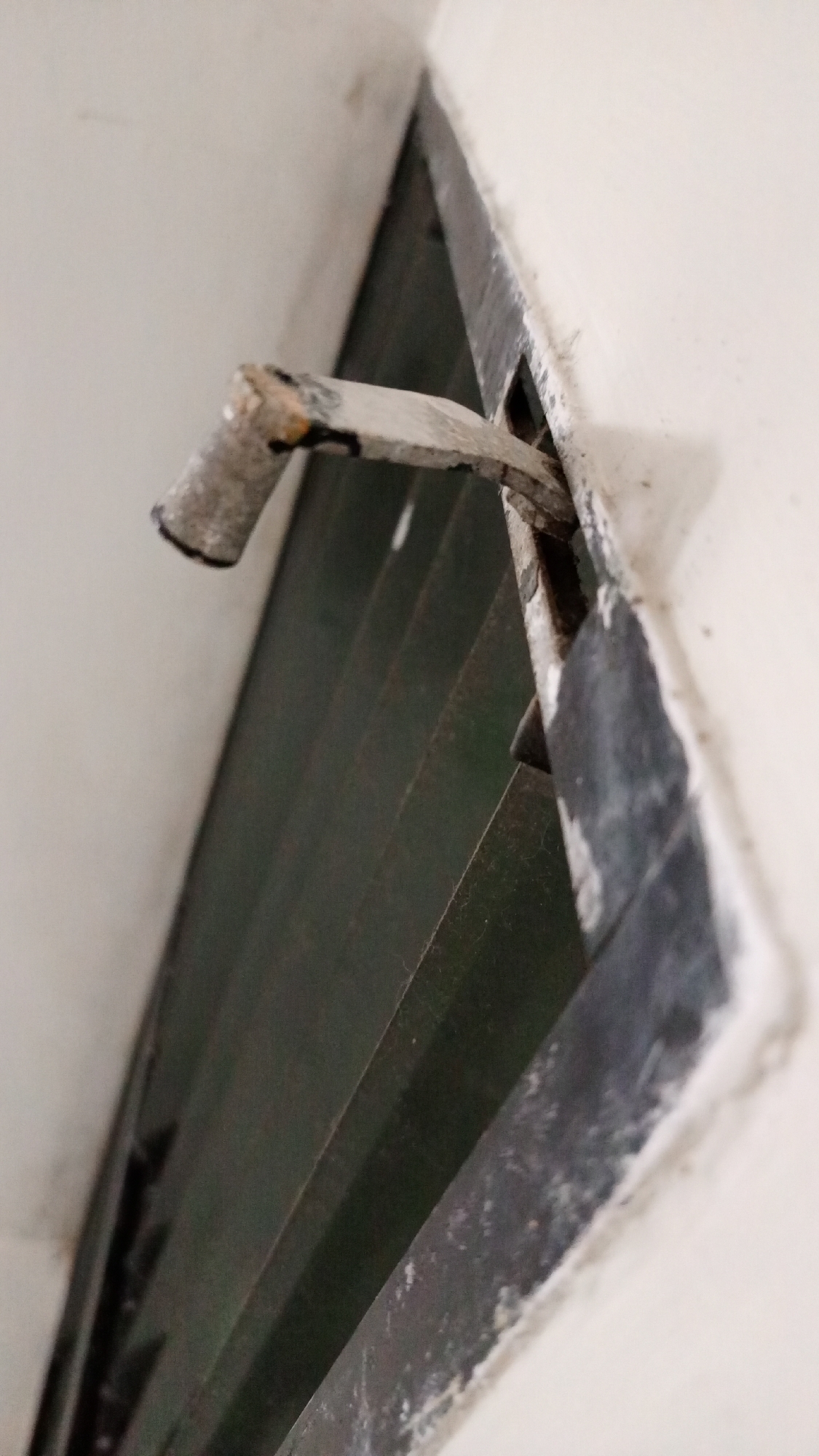 The image shows a close-up of an old, louvered window, possibly in a state of disrepair. The window frame is painted dark, with visible chipping and wear. A metal rod, possibly the control mechanism for the louvers, protrudes from the side of the window and appears bent or damaged. The louvers themselves are dark and appear slightly dusty. The surrounding wall is painted white and shows signs of wear and age. The location is Los Robles, Venezuela. Specific details beyond the window itself are not discernible from the image. There is no other person or subject in this Image.