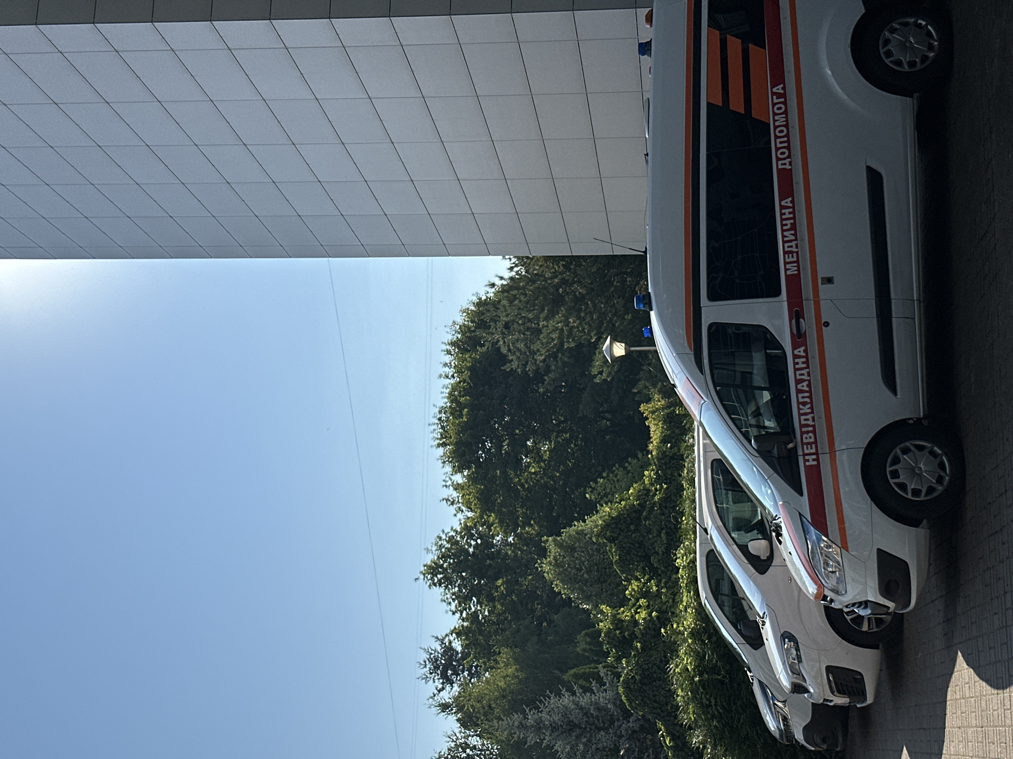 In the city of Kyiv, Ukraine, on a sunny day, several ambulances are parked in a row in front of a modern, tiled building. The closest ambulance, a white van with red and orange stripes, has the Ukrainian text "НЕВІДКЛАДНА МЕДИЧНА ДОПОМОГА" (Emergency Medical Assistance) printed on its side. Blue flashing lights are visible on the roof of this ambulance, as well as on another ambulance further back. Behind the ambulances, lush green trees and foliage form a dense natural backdrop. The sky is clear and blue.