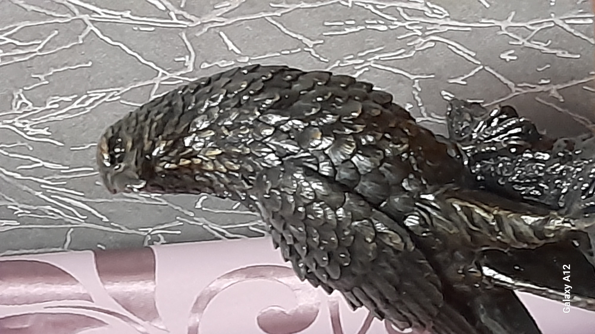 The image depicts an indoor scene in the city of Марковский, Russia. The central subject is a dark, metallic sculpture of a bird of prey, likely an eagle or hawk, positioned in profile and facing left. The sculpture exhibits detailed, overlapping feather textures, with glints of light reflecting off its surface, suggesting a bronze or dark metal material. It is perched on a dark, textured base resembling a tree branch or rock.
In the background to the left, a soft pink or light purple fabric with a large, abstract, swirling pattern is visible, likely part of a curtain or upholstered item. To the right of the sculpture, a light gray surface, possibly a wall, features an intricate white or light silver abstract pattern composed of thin, linear, branch-like elements. The bottom left corner of the image contains the visible watermark "Galaxy A12." No people or active interactions are present in the static scene.