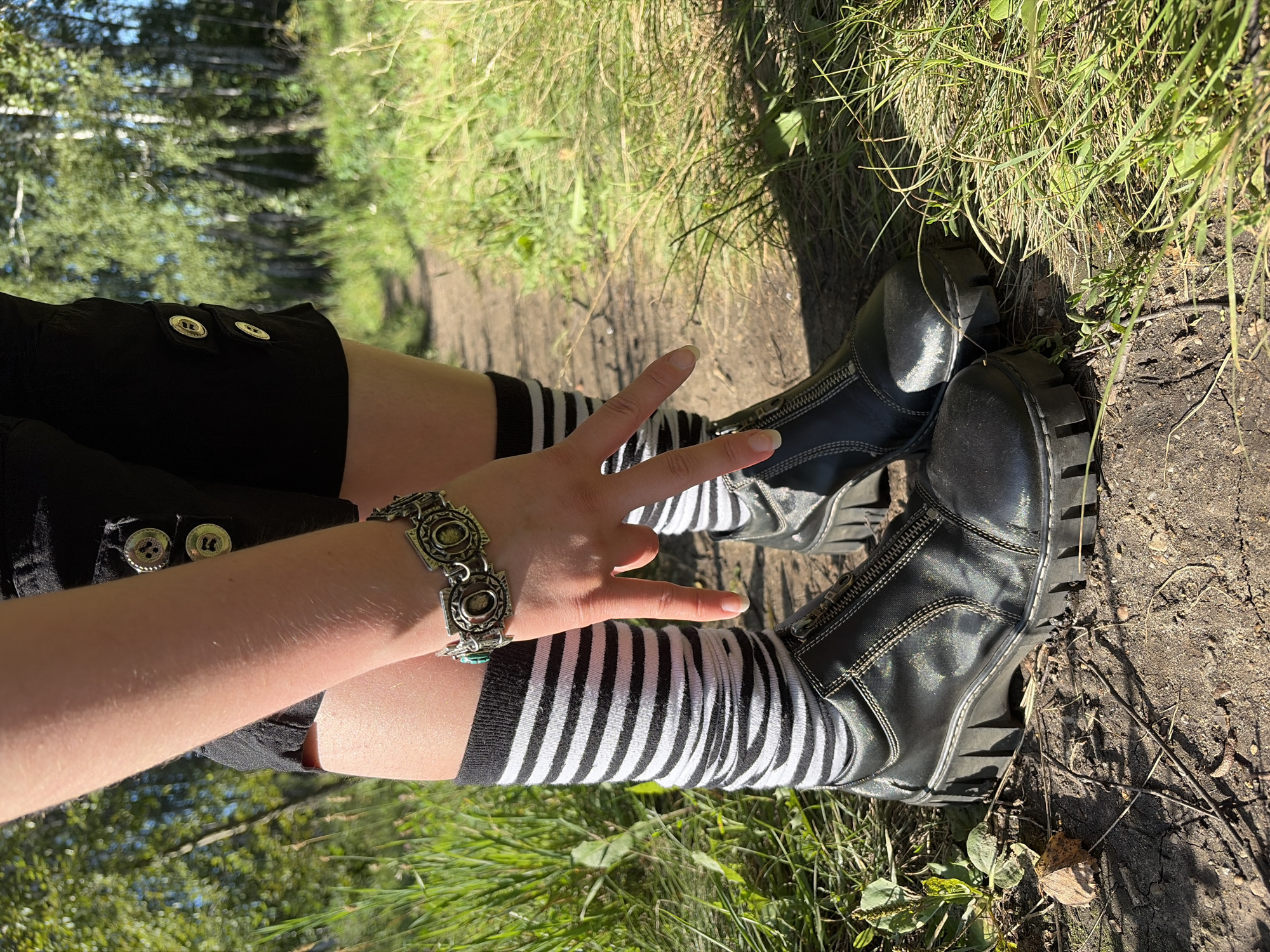 The image shows the lower legs and feet of a person wearing black shorts with two gold buttons on each side, black and white striped socks, and black zip-up boots with thick soles. The person is wearing a metal bracelet on their left wrist. Their left hand is held up, with the index and middle fingers extended in a V shape, with the index finger pointing downwards. The person is standing on a dirt path in a grassy, wooded area. The lighting suggests it is daytime. The image does not provide any specific details that would allow for localization to Chelyabinsk, Russia.