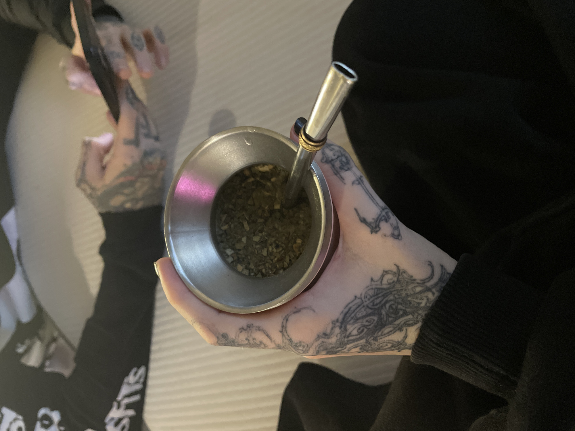 A person with tattooed hands holds a silver mate gourd with a metal bombilla, filled with yerba mate. Another person with tattoos on their hands holds a smartphone. The clothing is dark, and the background appears to be a light-colored surface. The photo depicts the act of drinking yerba mate, a common cultural practice. There is no readily discernible information to determine if this photo was taken in Buenos Aires, Argentina.