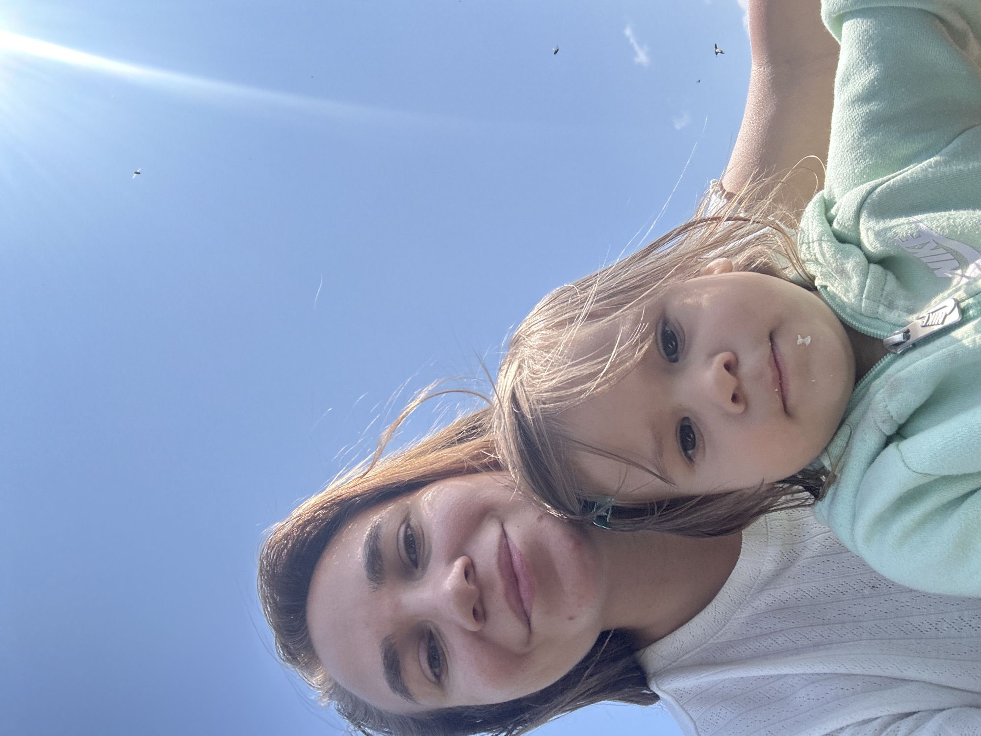 This is a selfie taken by two people, an adult and a child, against a bright blue sky with the sun shining. The adult, likely a woman, is in the foreground, smiling at the camera with her head tilted. The child, a young girl with blonde hair, is positioned above the adult, looking directly at the camera with a curious expression. The child is wearing a light green hooded jacket. The scene appears to be outdoors, possibly in a natural setting given the clear sky and distant objects that could be birds or insects. The lighting suggests it is daytime, likely mid-morning or afternoon, with the sun casting a strong glare. There are some small dark specks visible in the sky, which could be birds or insects, and a thin white contrail from an aircraft. The overall mood is happy and relaxed, captured in a casual, candid moment. The image is oriented sideways. There is visible text on the child's jacket, a white Nike logo on the zipper and sleeve.