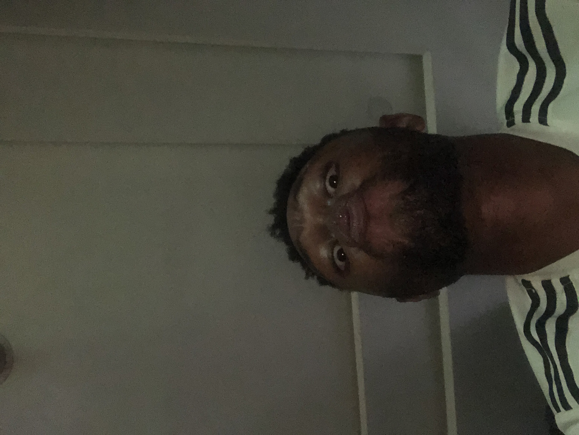 A close-up, vertically-oriented, low-light image shows a dark-skinned person with a beard, looking directly at the camera from a low angle. The person is wearing a white garment with black stripes on the right side of the frame. A neutral-colored wall is visible in the background. The location in the city is Unknown, Unknown.