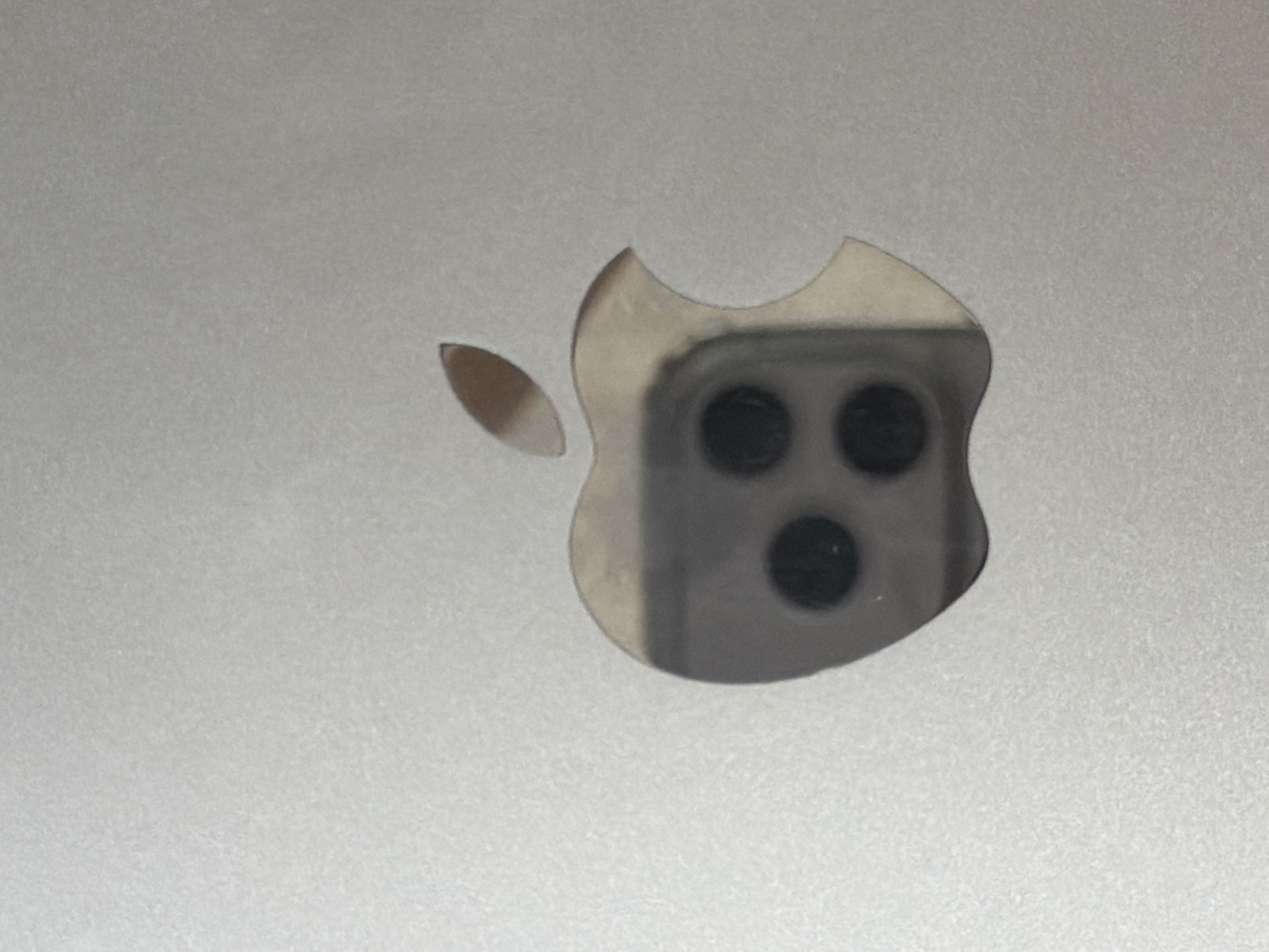 The image shows a close-up of the Apple logo, which appears to be engraved or cut out of a metallic surface. The Apple logo is silver and is reflecting a camera lens array that contains three circular black lenses. The surface around the logo is a textured, light gray metal. There is no indication of the city of Lyubimets, Bulgaria, in the image.