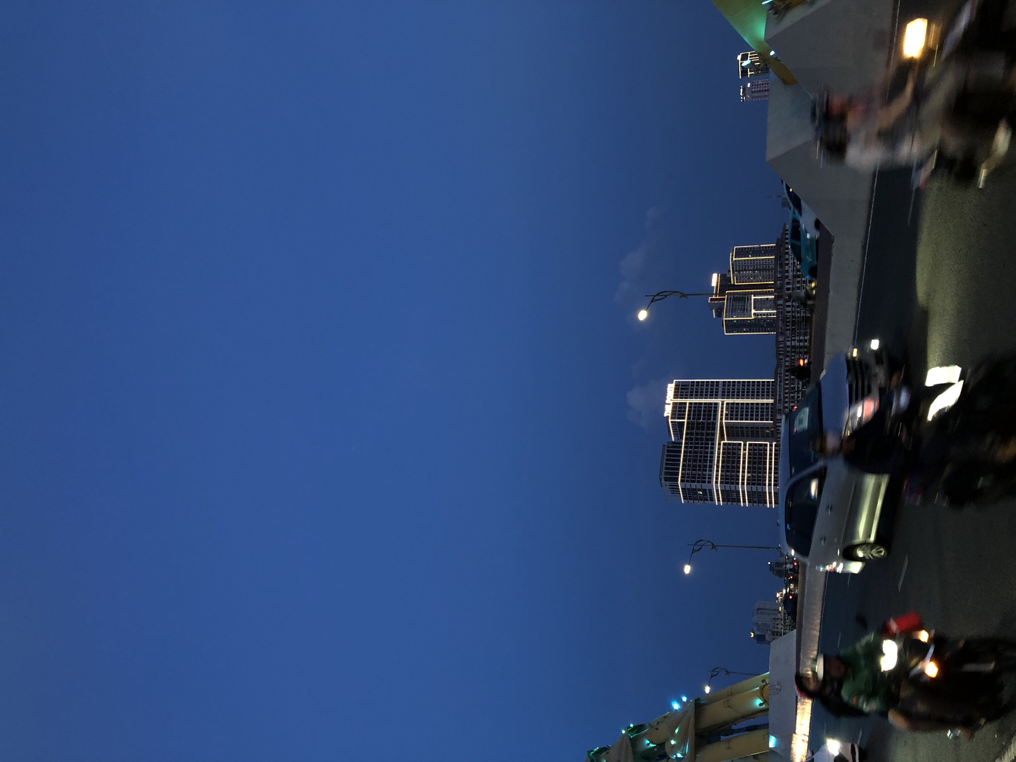 The image captures a nighttime scene on a bridge in Da Nang, Vietnam. A deep blue sky dominates the upper portion of the frame. In the middle ground, two illuminated modern buildings stand out, one with a distinctive "H" shape formed by its lights. To the left, a bridge structure is visible, adorned with green and blue lights. The foreground is a busy roadway on the bridge, with several motorcycles in motion, their headlights and taillights creating streaks of light due to the long exposure. One motorcycle in the center carries two people. Another motorcycle on the right is also visible with its lights. A silver minivan is also present on the bridge. The overall impression is a bustling urban environment at dusk or night.
