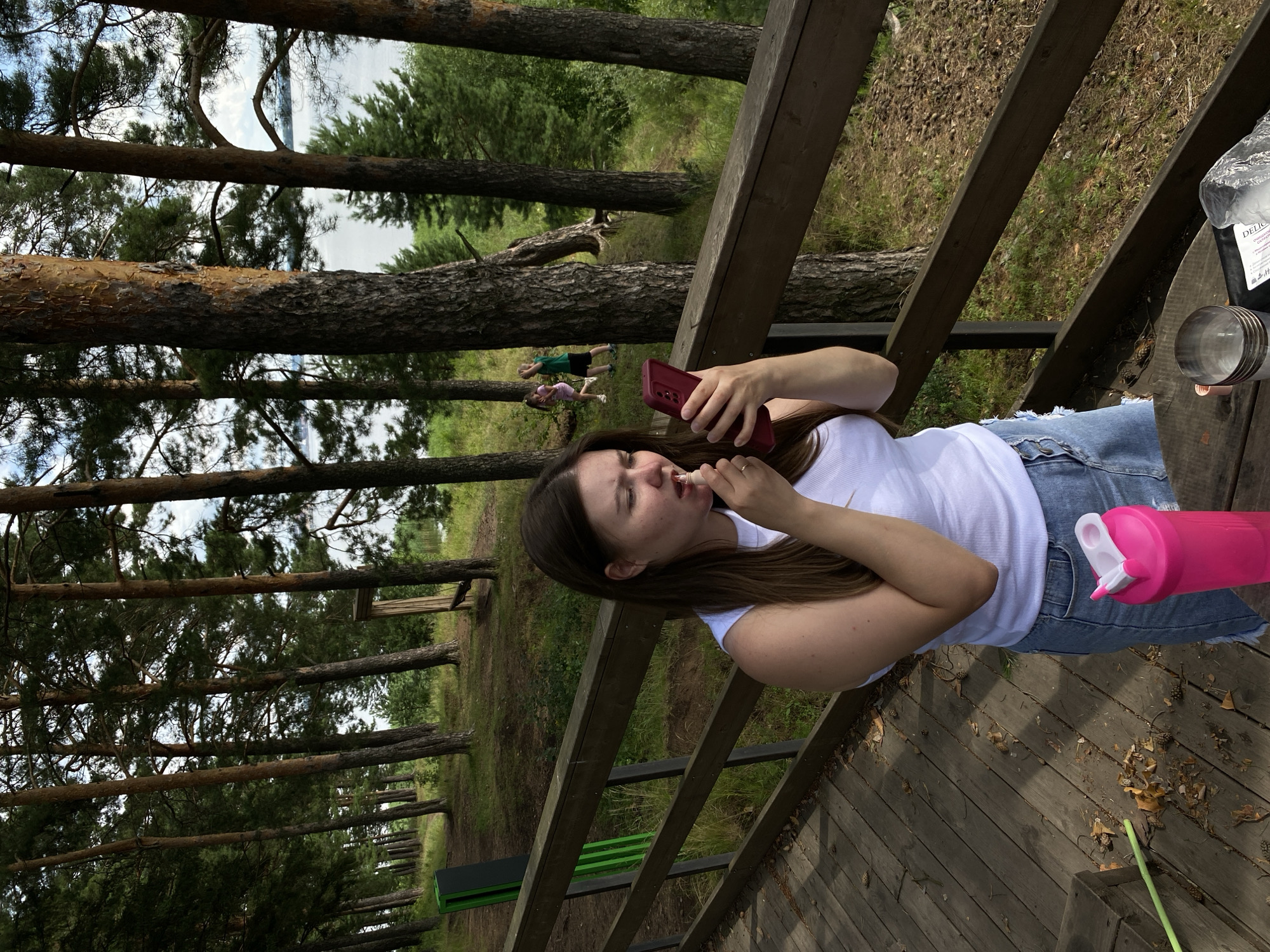 A young woman with long, brown hair is applying lipstick while looking at her red smartphone. She is wearing a white sleeveless top and a denim skirt. She is standing on a wooden deck with a wooden railing in front of her. To her right, on a wooden table, is a pink shaker bottle, a glass, and a plastic bag. In the background, there are tall pine trees, green grass, and a body of water. Two children are visible in the distance playing. The scene appears to be a public outdoor area, possibly a park or recreational facility, located in Votkinsk, Russia.