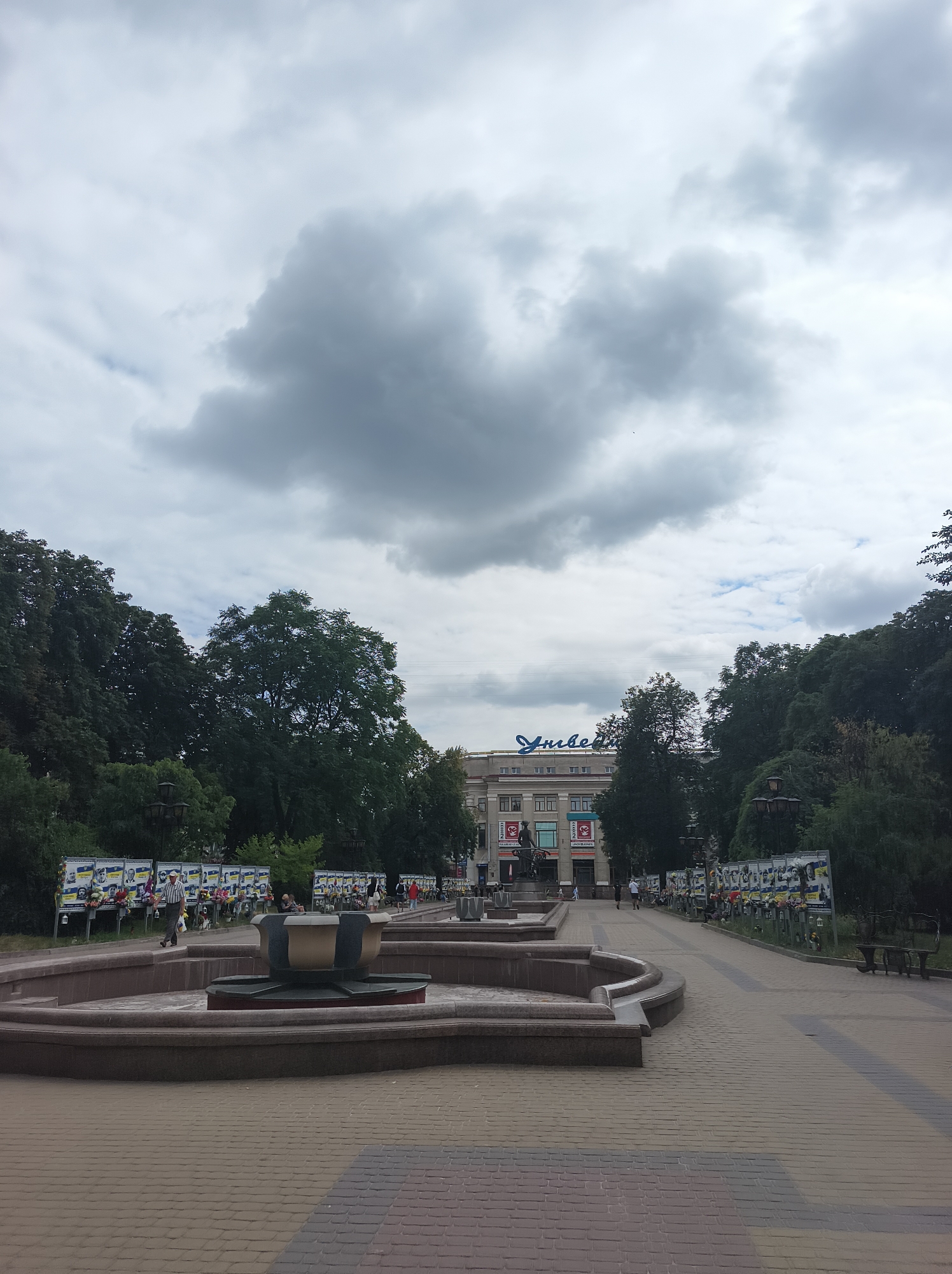 The image depicts a wide, paved pedestrian walkway in front of a fountain in Ternopil, Ukraine. The fountain, made of stone, is a prominent feature in the foreground, with a layered design and a central decorative element. The walkway is paved with interlocking bricks and is lined with lampposts and benches. Along the left side of the walkway, a series of display boards with portraits and Ukrainian flags are arranged. These appear to be memorials. In the background, a large building with the blue Cyrillic lettering "Універмаг" (Univermag, meaning department store) is visible, along with trees and power lines. Several people are walking along the path, some in the distance. The sky is overcast with dark clouds.