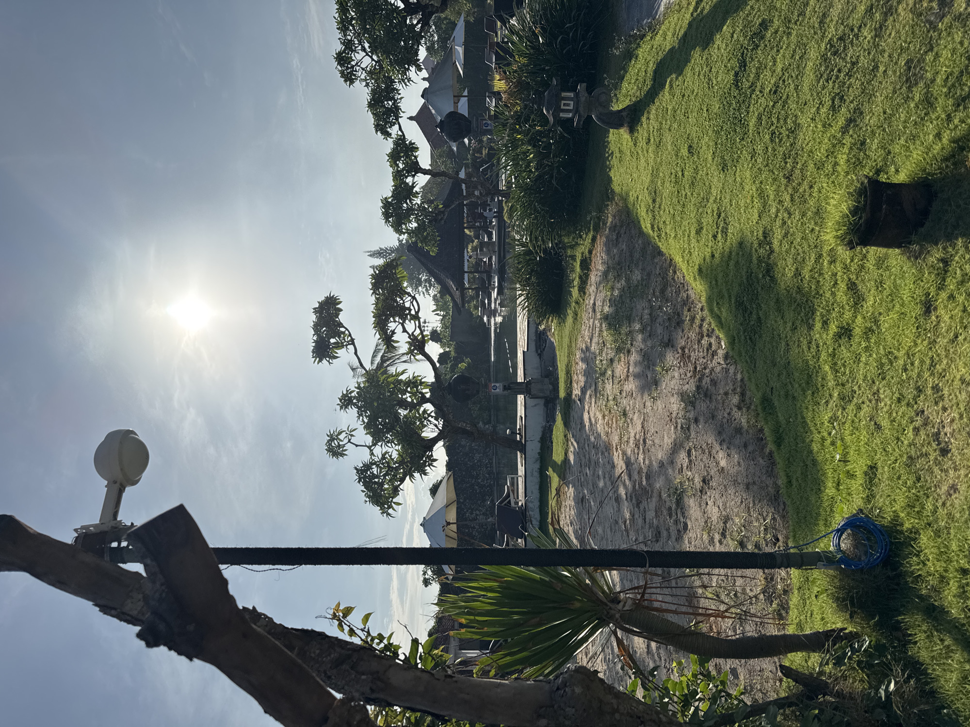 The scene is an outdoor area in Jimbaran, Indonesia. The sun is high in the sky, creating bright conditions. The foreground features a tree branch with a white, dome-shaped device attached to it. A black pole runs vertically near the tree. A coiled blue cable sits on the green grass adjacent to a sandy path.
In the background, a thatched-roof structure and umbrellas suggest a recreational or resort area. There are trees, shrubs, and a grassy lawn. The location appears to be a well-maintained beachfront or garden setting.