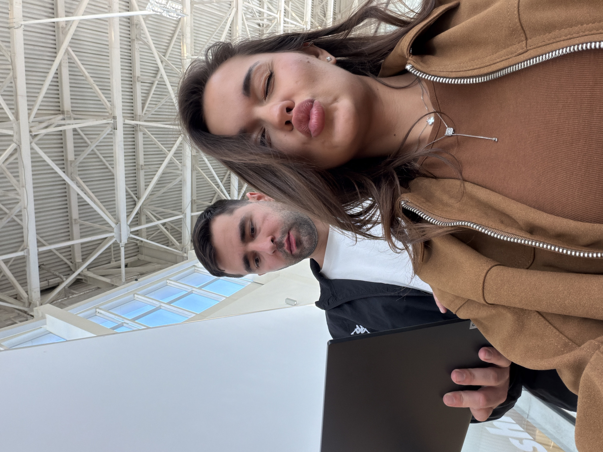 The image is a portrait-oriented selfie, rotated 90 degrees counter-clockwise. It features two individuals, a woman and a man, captured indoors under a large, modern ceiling.
The woman is in the foreground, positioned on the right side of the rotated image. She has long, dark hair and is making a playful "kiss face" with pursed lips and closed eyes. She is wearing a brown zippered jacket over a brown top. Two silver necklaces adorn her neck: one with a four-leaf clover pendant and another longer chain with a square pendant. A diamond stud earring is visible in her ear.
Behind and to the left of the woman is a man with dark hair and a beard. He wears a white t-shirt under a black jacket, which features a white Kappa logo on the left arm. He is holding a black laptop and appears to be looking towards the camera with a neutral expression.
The background shows a spacious indoor setting with a high, architecturally distinct ceiling. The ceiling is composed of intricate white metal trusses and beams. Several skylights are visible within the ceiling structure, allowing natural light to enter and showcasing a clear blue sky, indicating it is daytime. The overall setting suggests a large public space such as an airport terminal, a modern shopping center, or a convention hall. The mood is casual and playful. The location context is Krasnoyarsk, Russia.