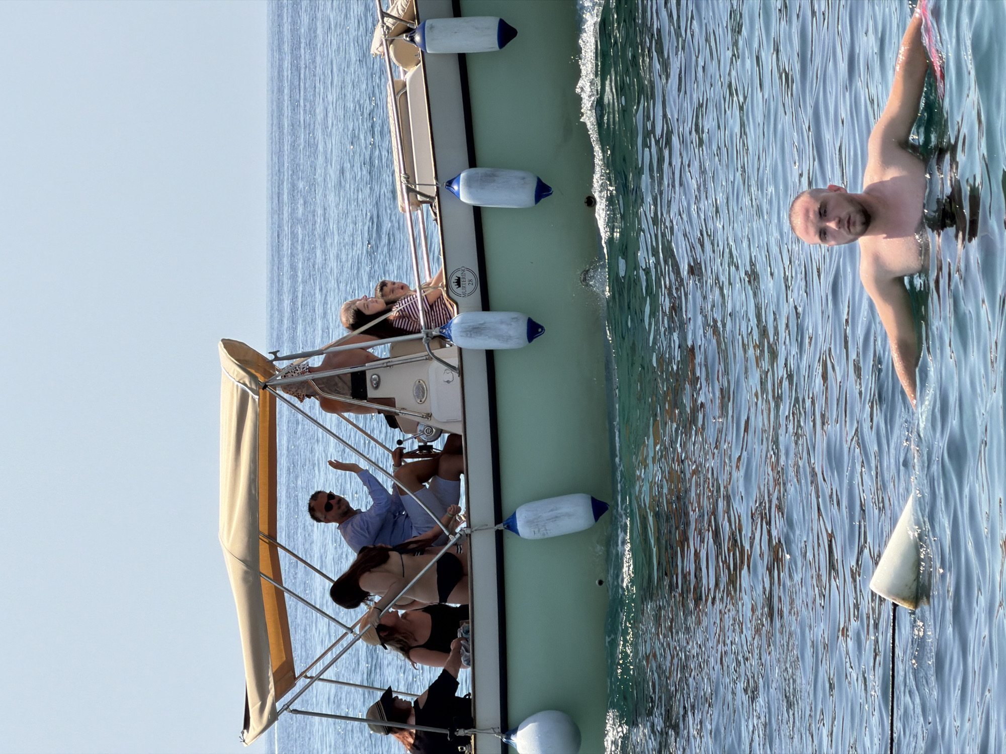A man swims in the water in the foreground, facing the camera with his arms outstretched. Behind him, a boat with several people on it sails on the water. The boat has a light-colored canopy and is painted pale green. People are seated and standing on the boat. The ocean is calm and blue under a clear sky. The scene is likely on the coast of Budva, Montenegro, given the context.