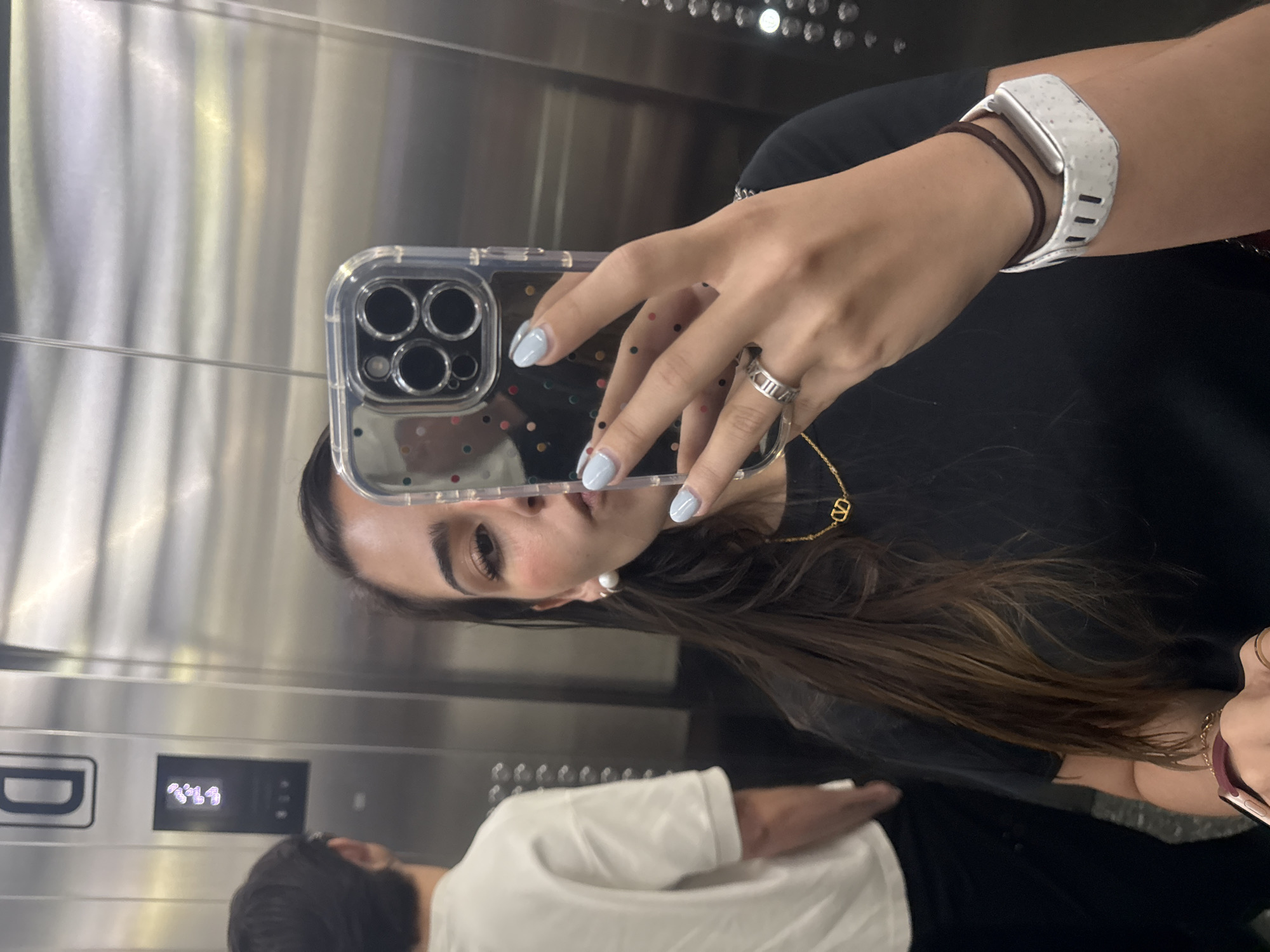 A person is taking a selfie in what appears to be an elevator. The person is holding a smartphone with a clear case, and their face is partially visible in the reflection. The person has manicured nails painted light blue and is wearing a silver ring, a gold necklace with a "V" pendant, and a white and brown wristband. In the background, another person wearing a white shirt is visible in the elevator's mirrored wall. The interior of the elevator is stainless steel. The location is not possible to determine given the available information.