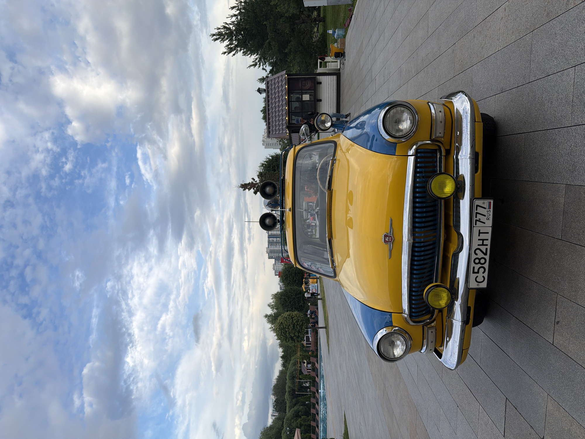 An eye-level, medium shot captures a vintage yellow and blue car parked on a paved walkway with a cloudy sky above. The car is centrally positioned and faces the viewer directly. It has chrome accents, including the bumper, grille, and headlights. Two yellow fog lights are mounted below the headlights. A license plate with the alphanumeric characters "C582HT 777" and the Russian flag is affixed to the front. The car has two speakers mounted on its roof. To the left of the car, a swimming pool with lounge chairs is visible. To the right, a small, brown-roofed structure that appears to be a kiosk or food stand is present. In the background, there are trees, apartment buildings, and a few people walking. The overall lighting suggests it is late afternoon.