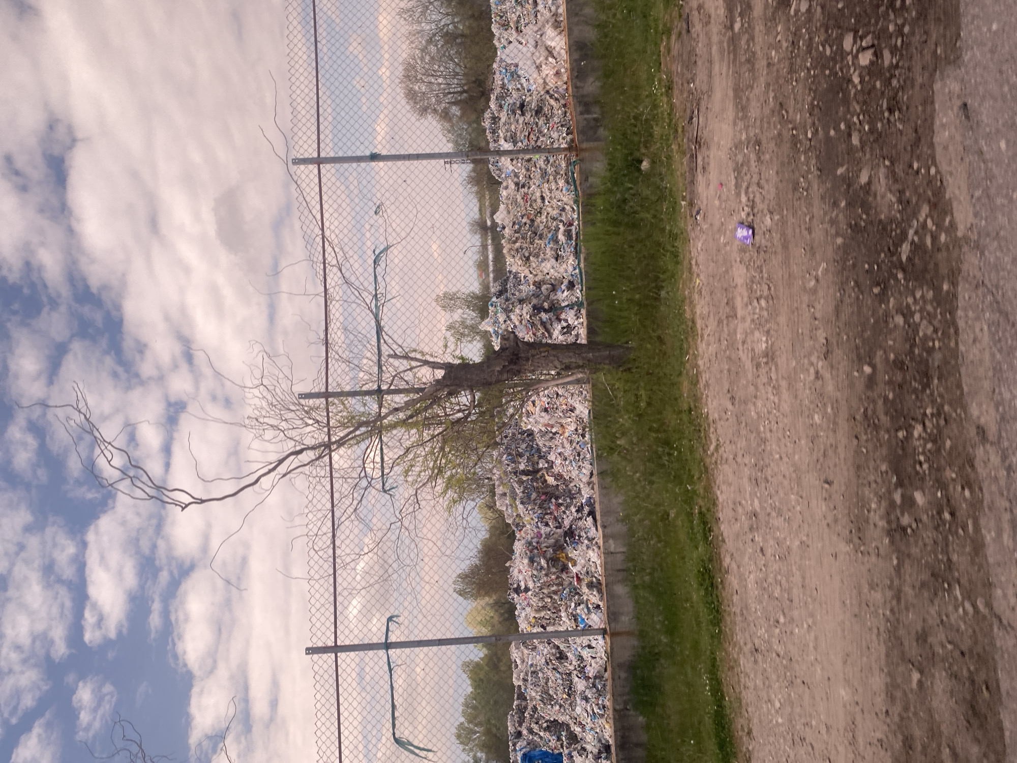 The image shows a landfill in what may be Kapatovo, Bulgaria. A chain-link fence separates a large pile of garbage from a narrow dirt road. A tree with bare branches stands next to the fence. The sky is visible behind the fence with scattered clouds. A small, purple object, potentially discarded trash, lies on the dirt road. There are no people visible in the image.