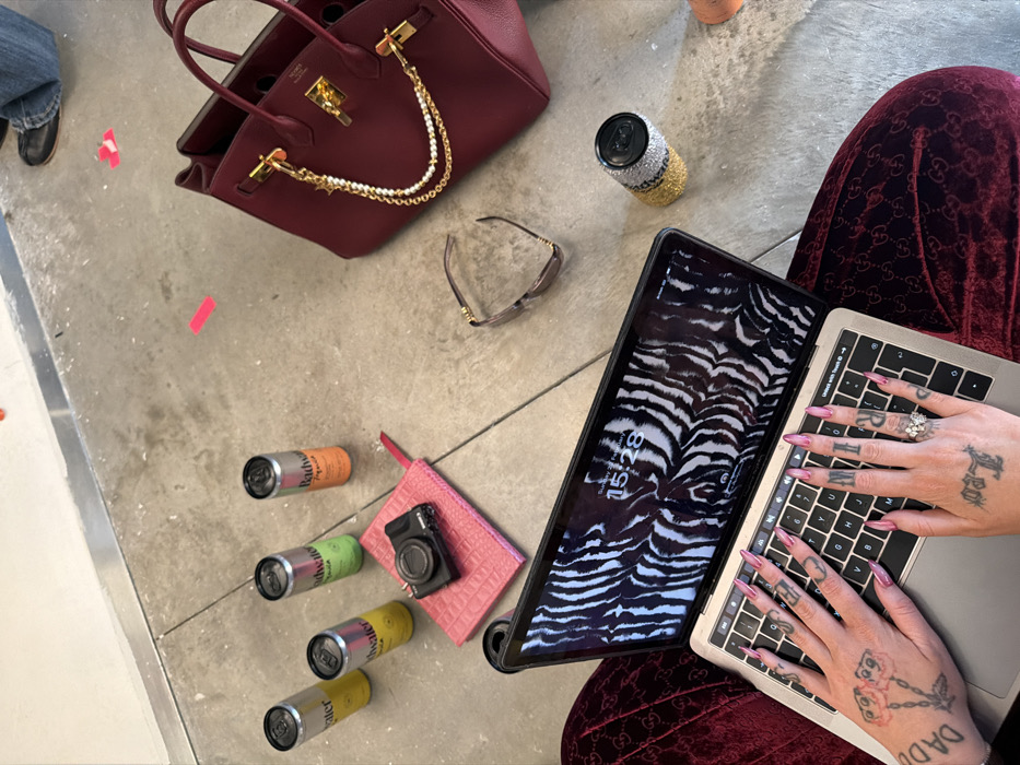 A burgundy handbag with gold hardware rests near a sparkling can. Sunglasses and several cans of drinks are scattered around a pink pouch and a black camera. A person sits with a laptop and a tablet displaying a zebra-striped screen. The person's hands, adorned with rings, tattoos, and long pink nails, are positioned on the laptop keyboard. The person wears burgundy velvet clothing.