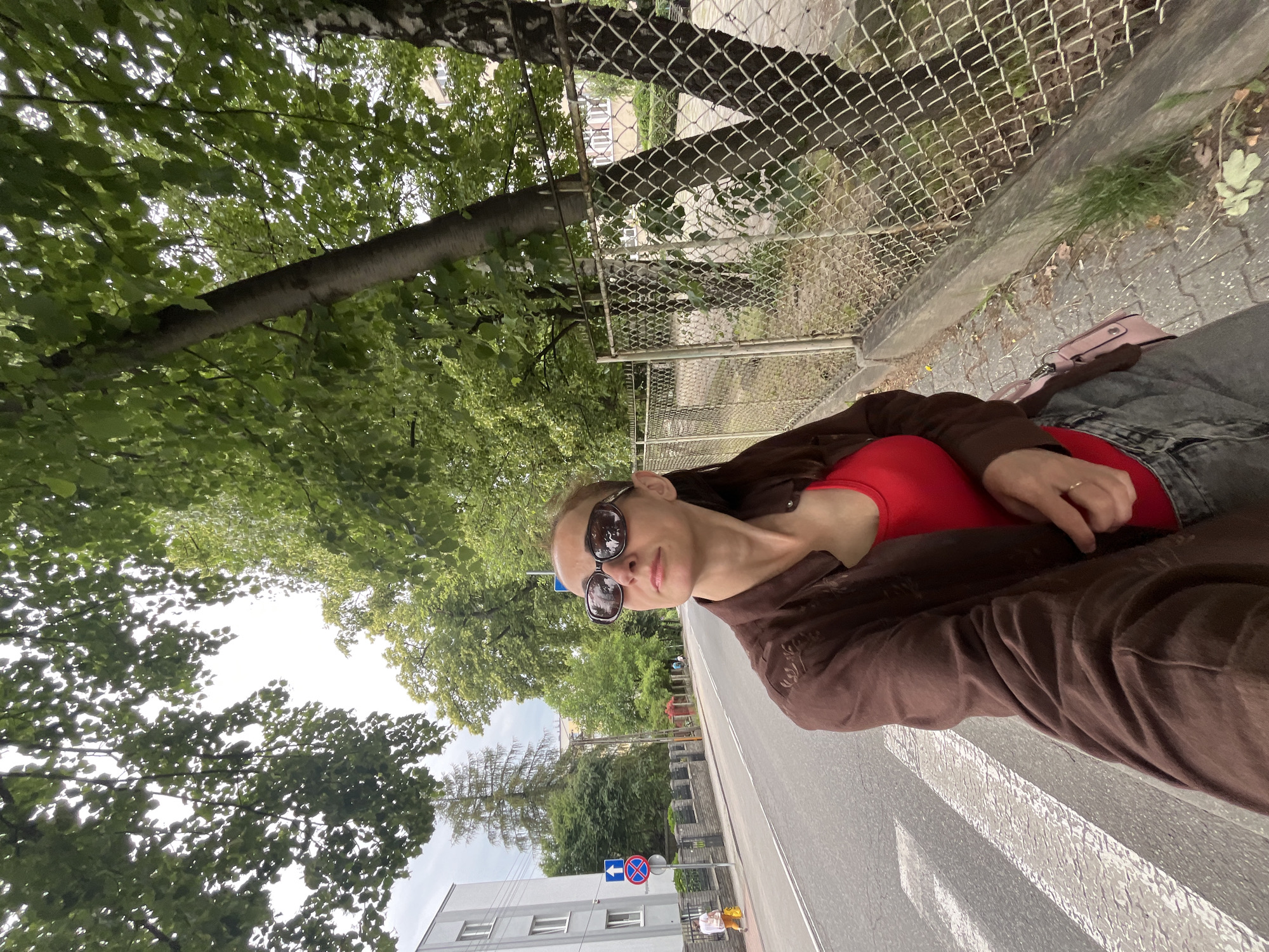 The image shows a young woman taking a selfie while sitting by the side of a road. She is wearing a red top and a brown jacket, and sunglasses. A pink purse is visible on her lap. The background features trees, a chain-link fence, and a road with a pedestrian crossing. The lighting suggests it is daytime, and the greenery indicates a temperate climate. The woman's expression is neutral, and she appears relaxed. There is no visible text in the image.